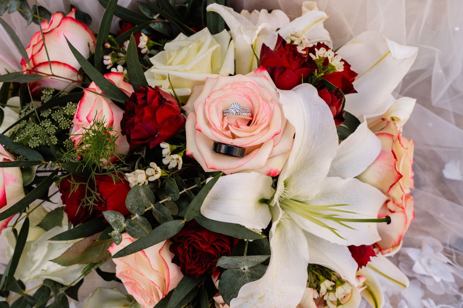 Elegant wedding bouquet featuring pink roses, red flowers, and white lilies with a diamond ring nestled among the blooms.