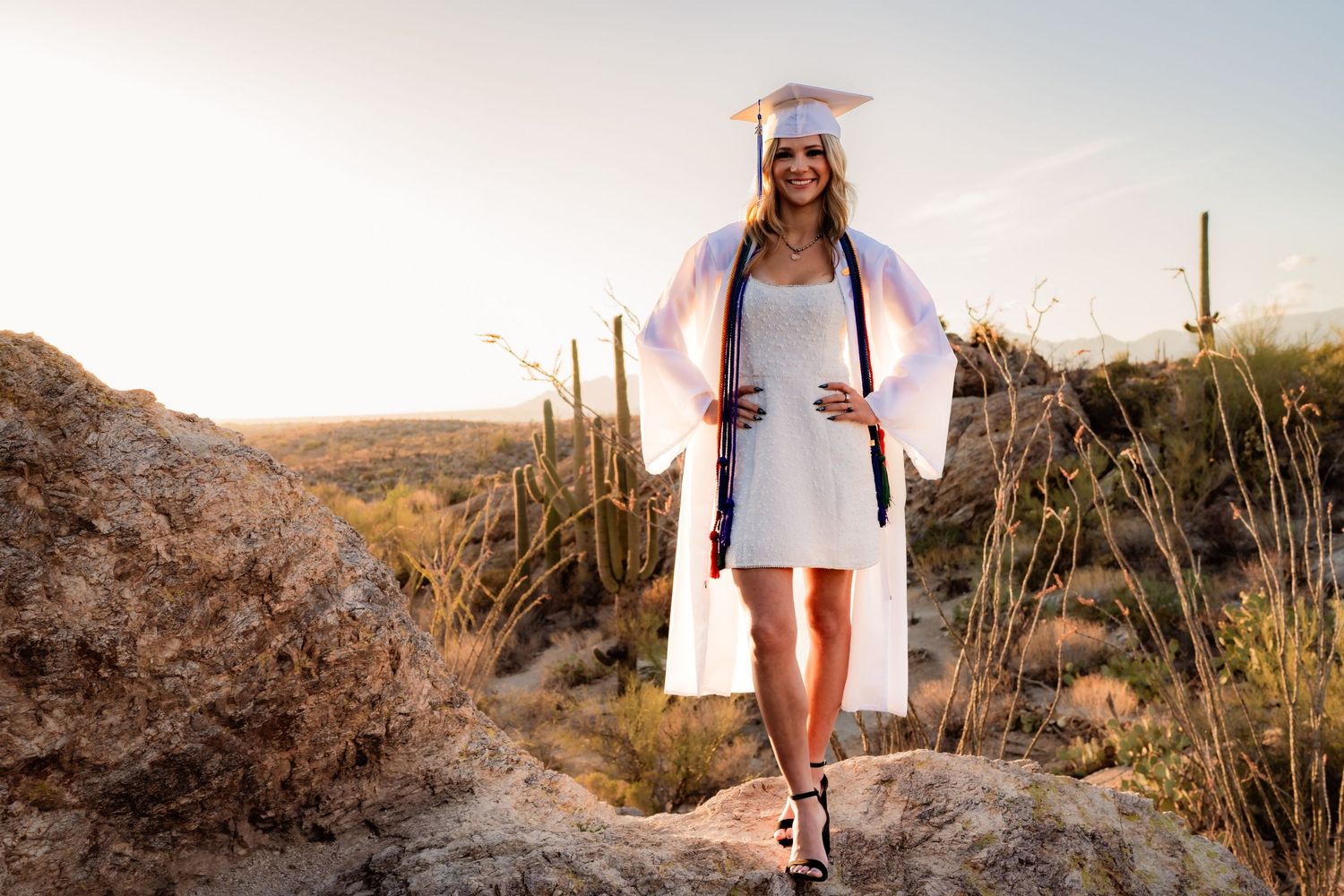 Young woman in white graduation cap and gown posing on desert rocks with saguaro cacti at sunset.
