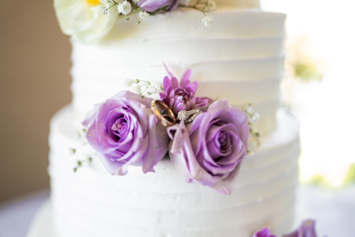 White tiered wedding cake decorated with purple roses and baby's breath flowers.
