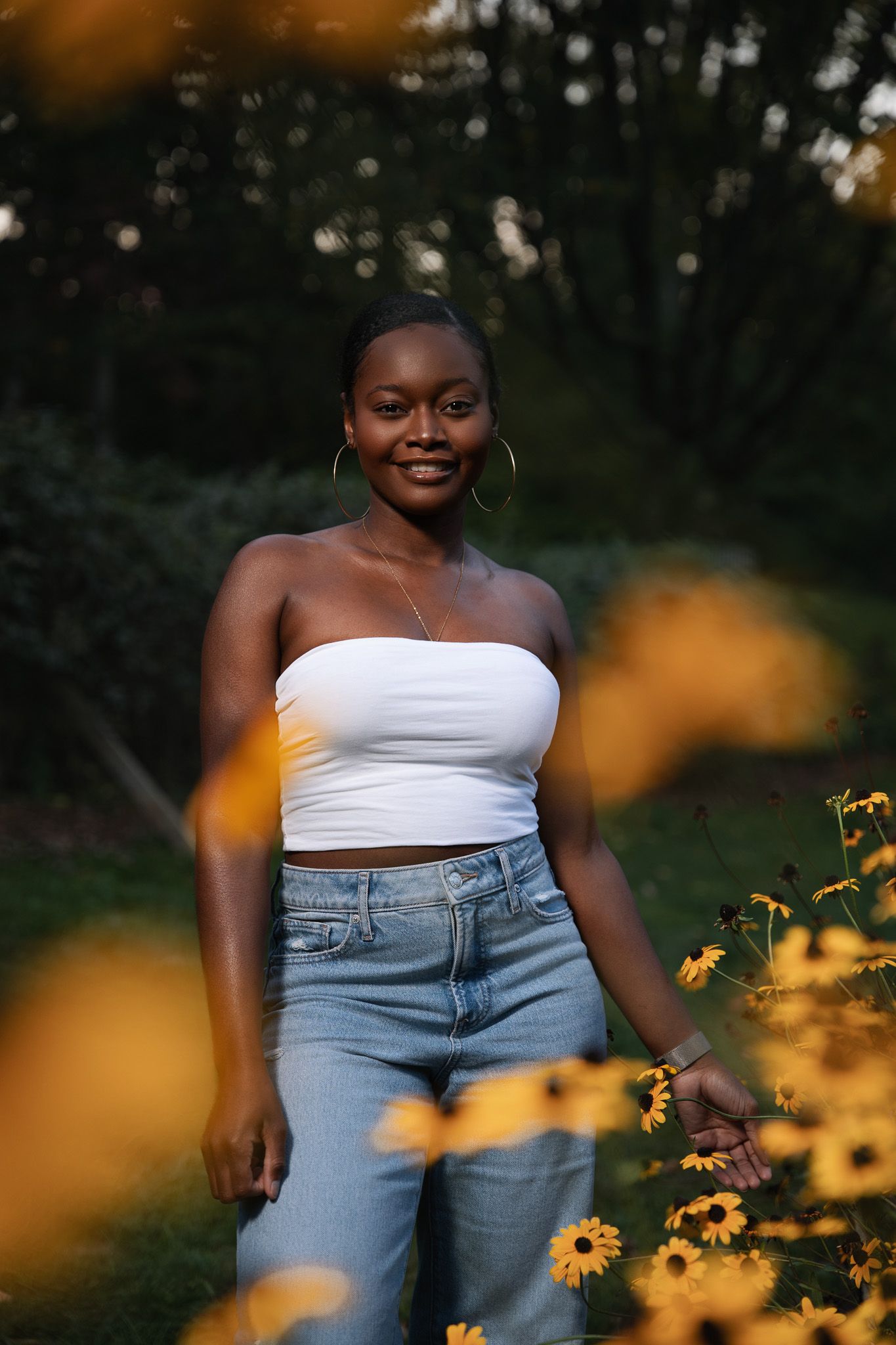 Smiling young Black woman in white tube top and jeans stands among yellow wildflowers in a garden portrait.
