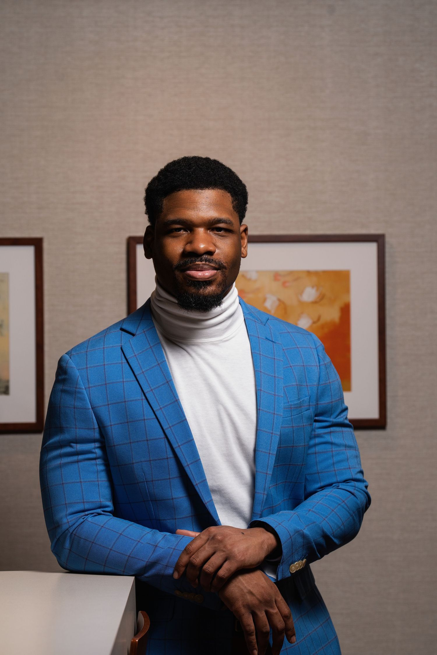 Smiling Black man in blue plaid blazer and white turtleneck leans against a table in a professional portrait.