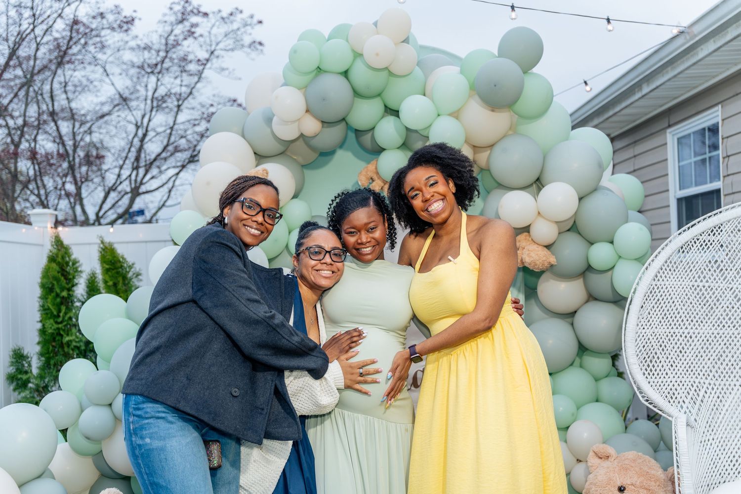 Four women smiling together at a baby shower with green and white balloon arch backdrop outdoors.