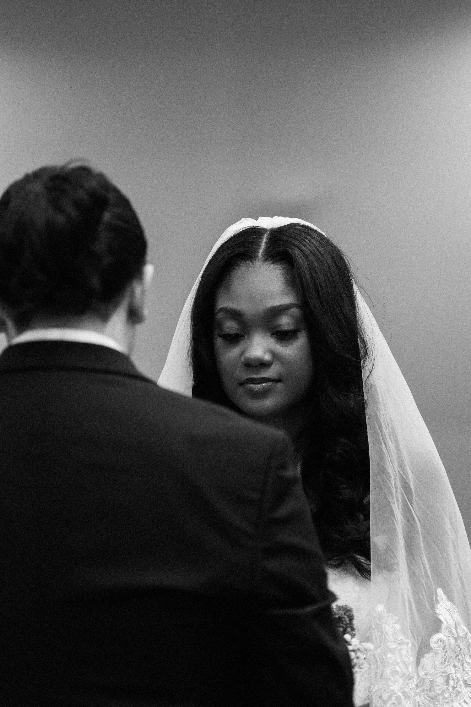 Black and white photo of bride in veil looking down during wedding ceremony with groom standing before her.