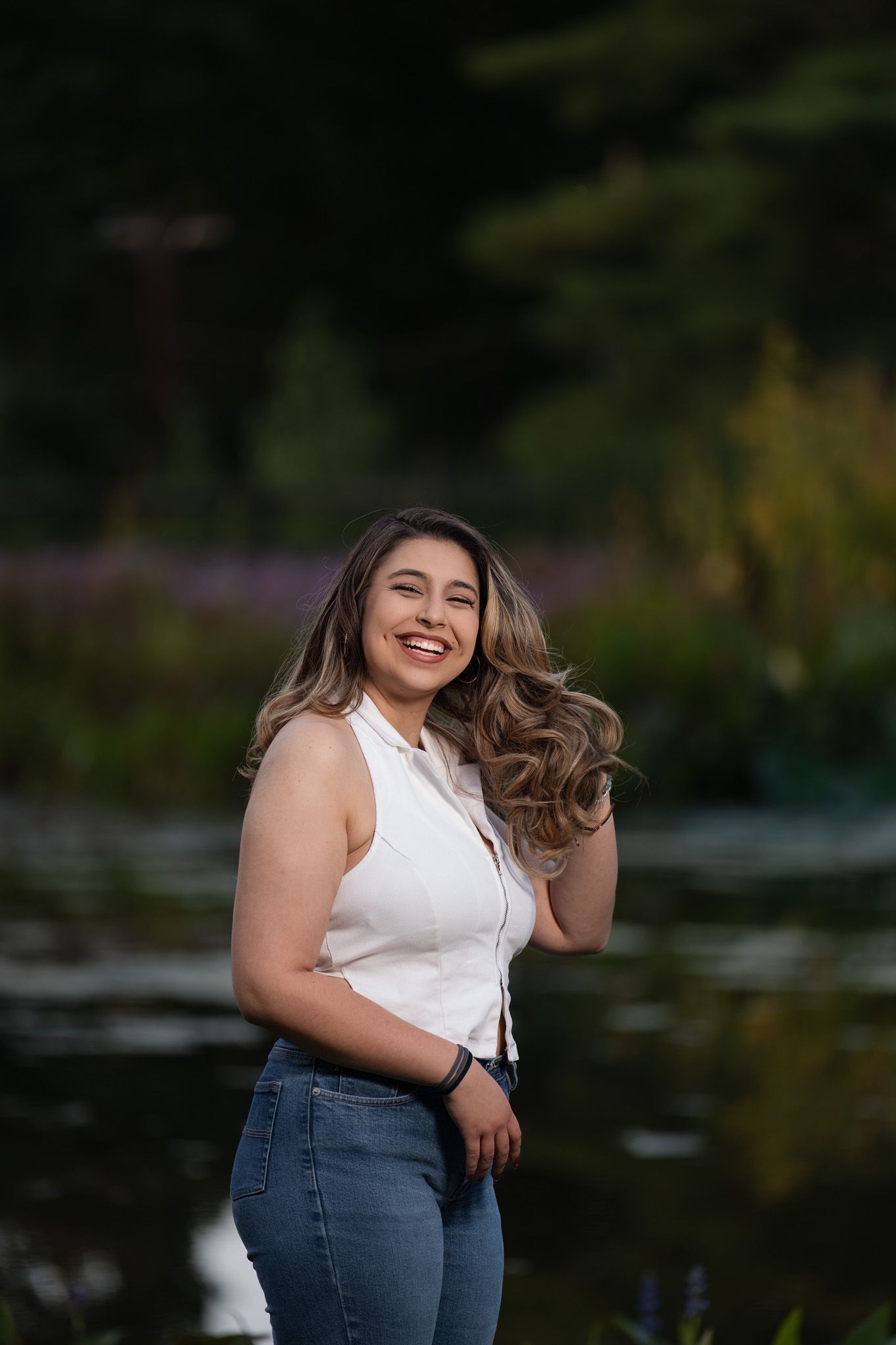 Smiling young woman in white top and jeans poses outdoors near a pond with lush green background.