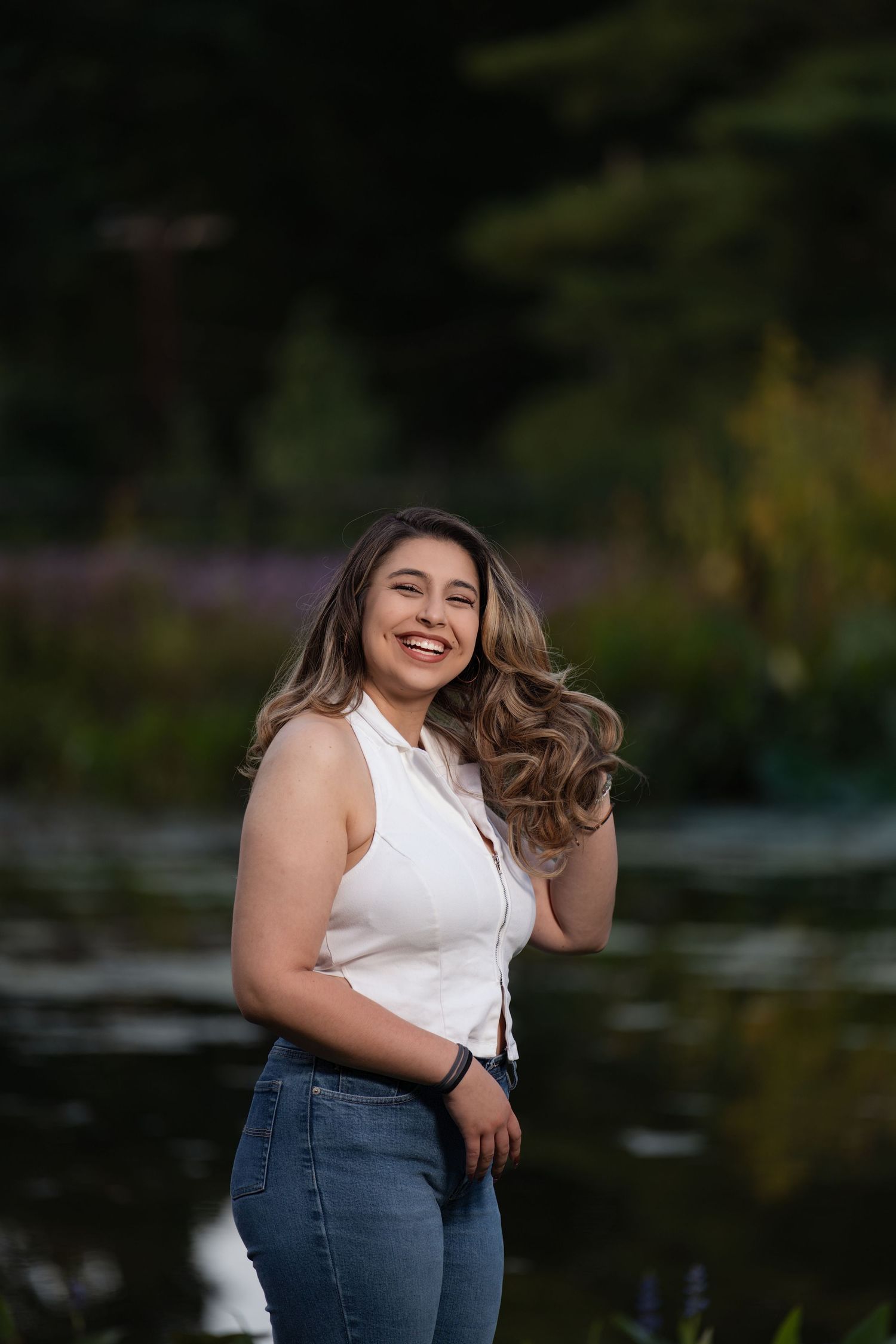 Young woman in white top and jeans smiling outdoors near a pond with lush greenery background.