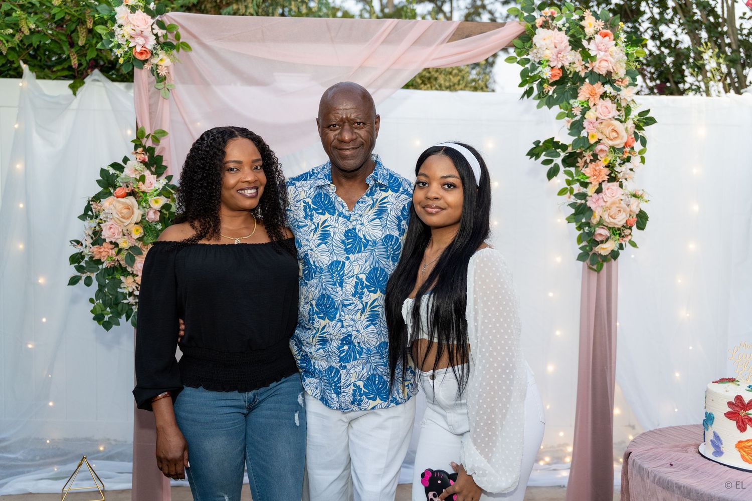 Man and two women smiling together in front of floral arch at outdoor garden party.