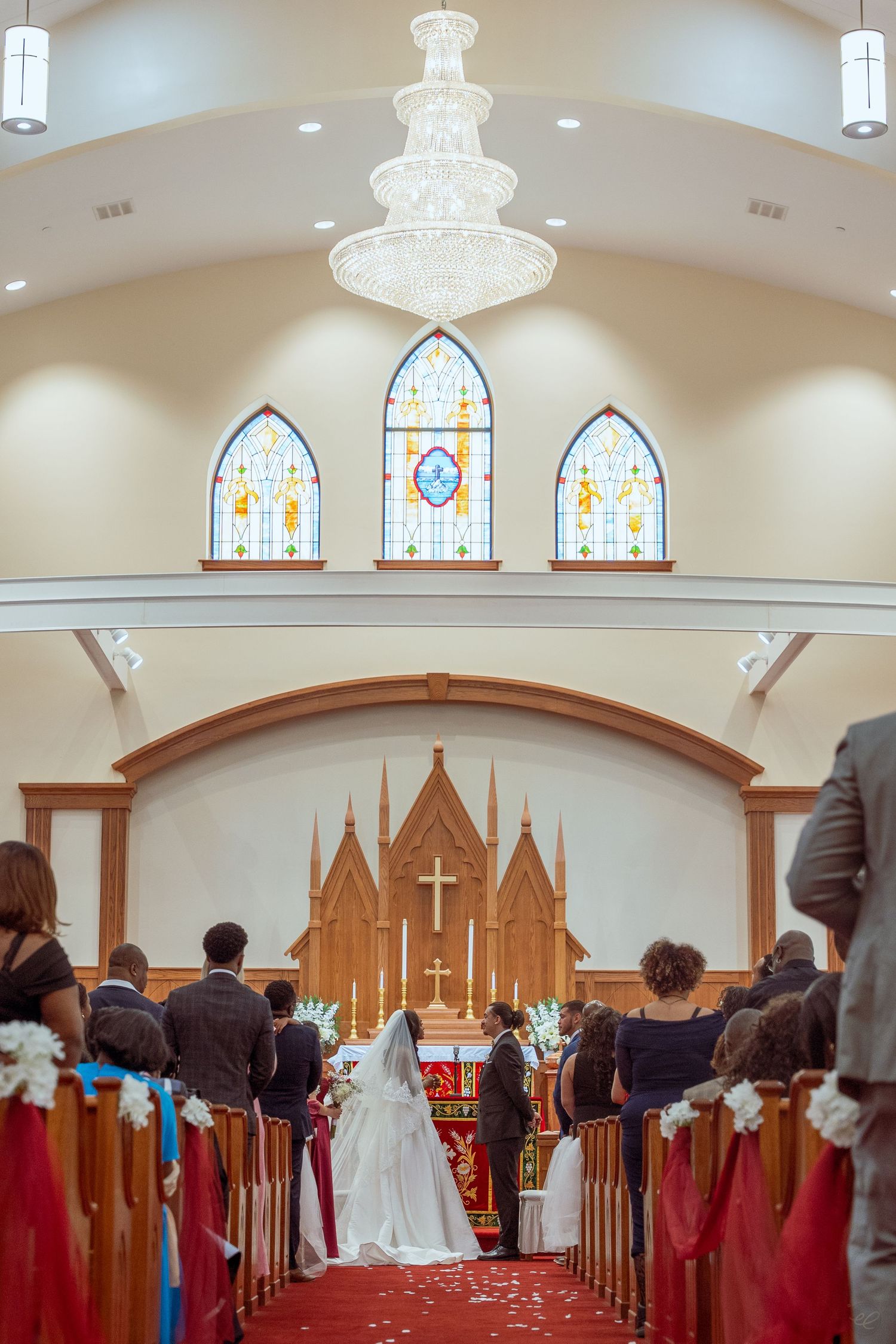 A wedding ceremony in a white chapel with stained glass windows, chandelier, and guests seated in wooden pews.