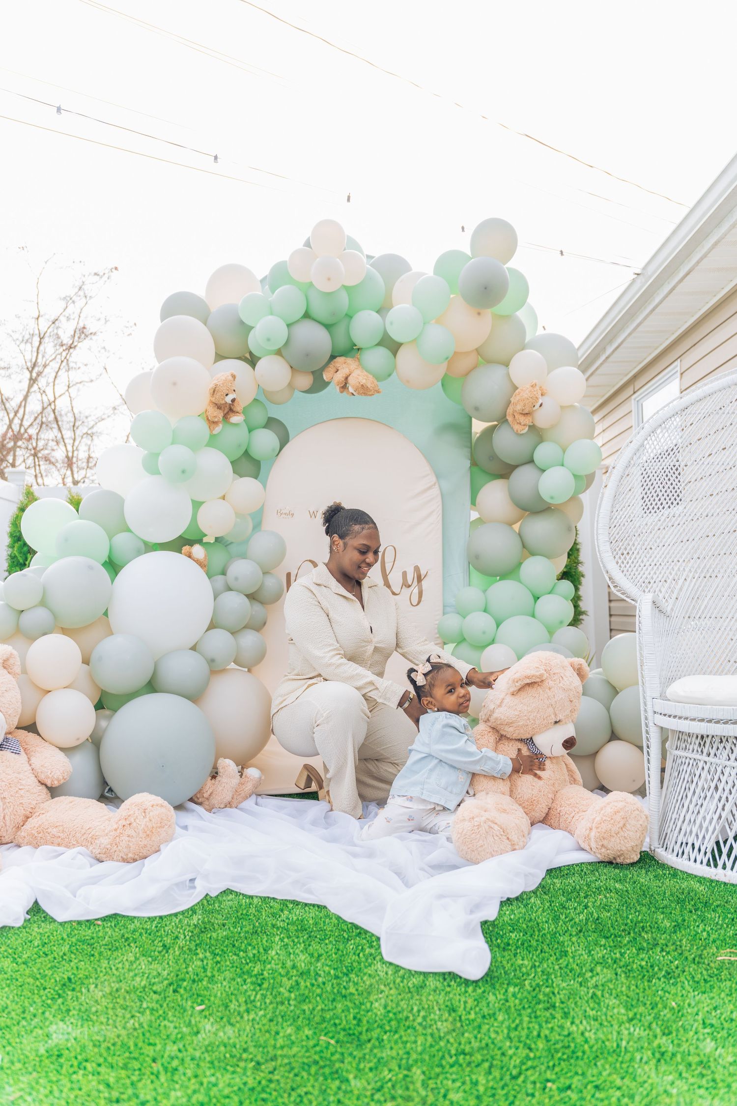 A woman sits with a toddler amid giant teddy bears under a mint green and gold balloon arch at an outdoor celebration.