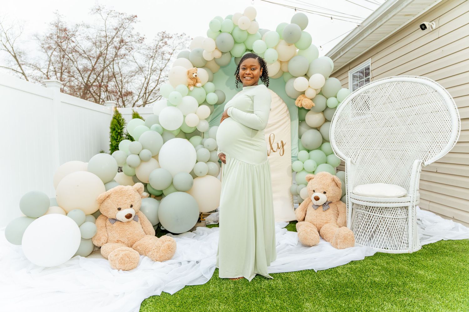 Pregnant woman in sage green gown poses at outdoor baby shower with balloon arch and giant teddy bears.