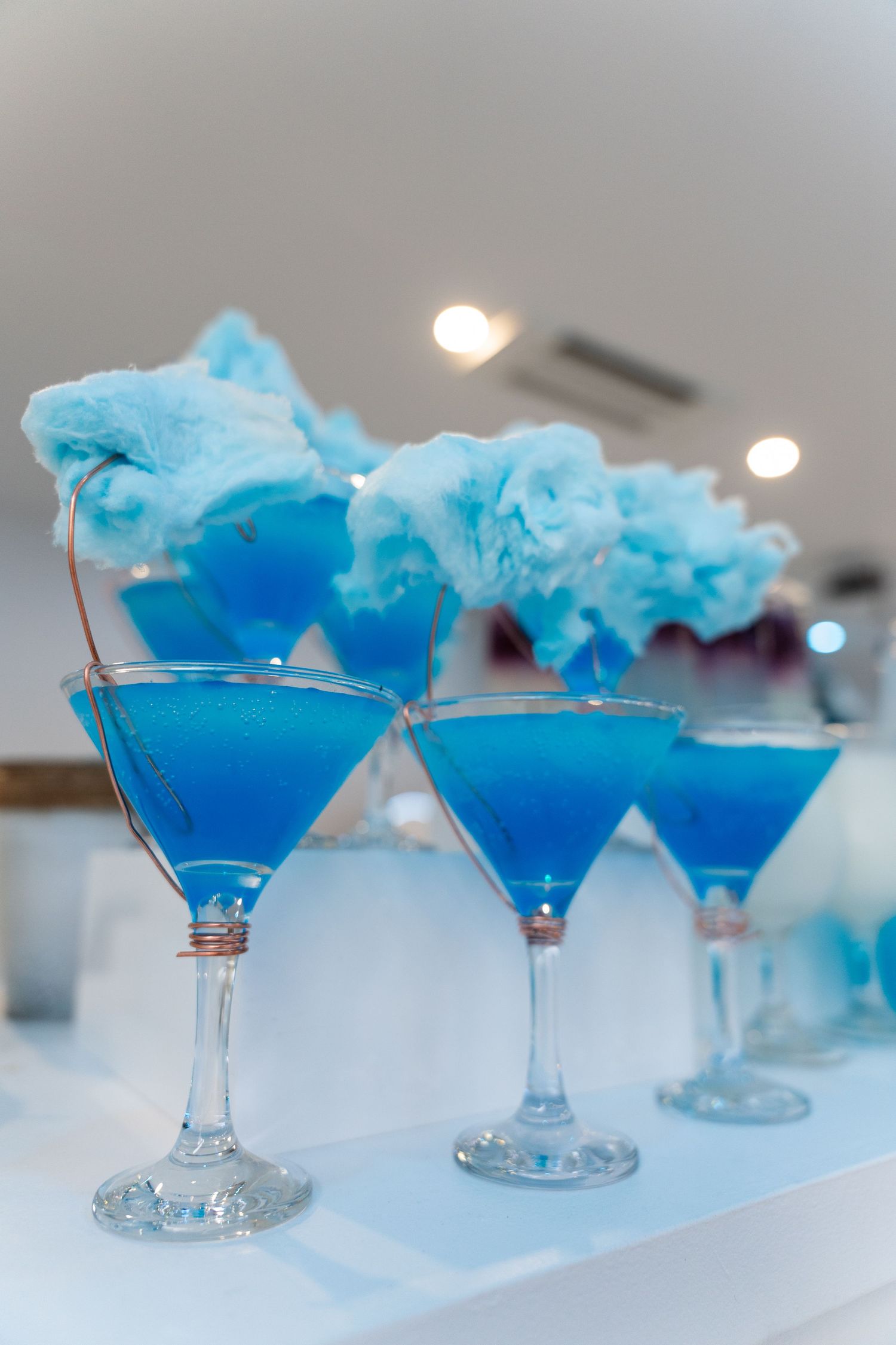 Blue cocktails in martini glasses topped with fluffy cotton candy displayed on a white bar counter.