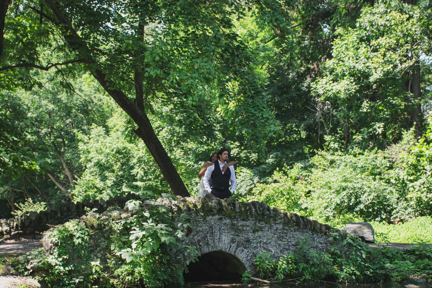 Person in formal attire standing on a rustic stone arch bridge surrounded by dense green foliage.