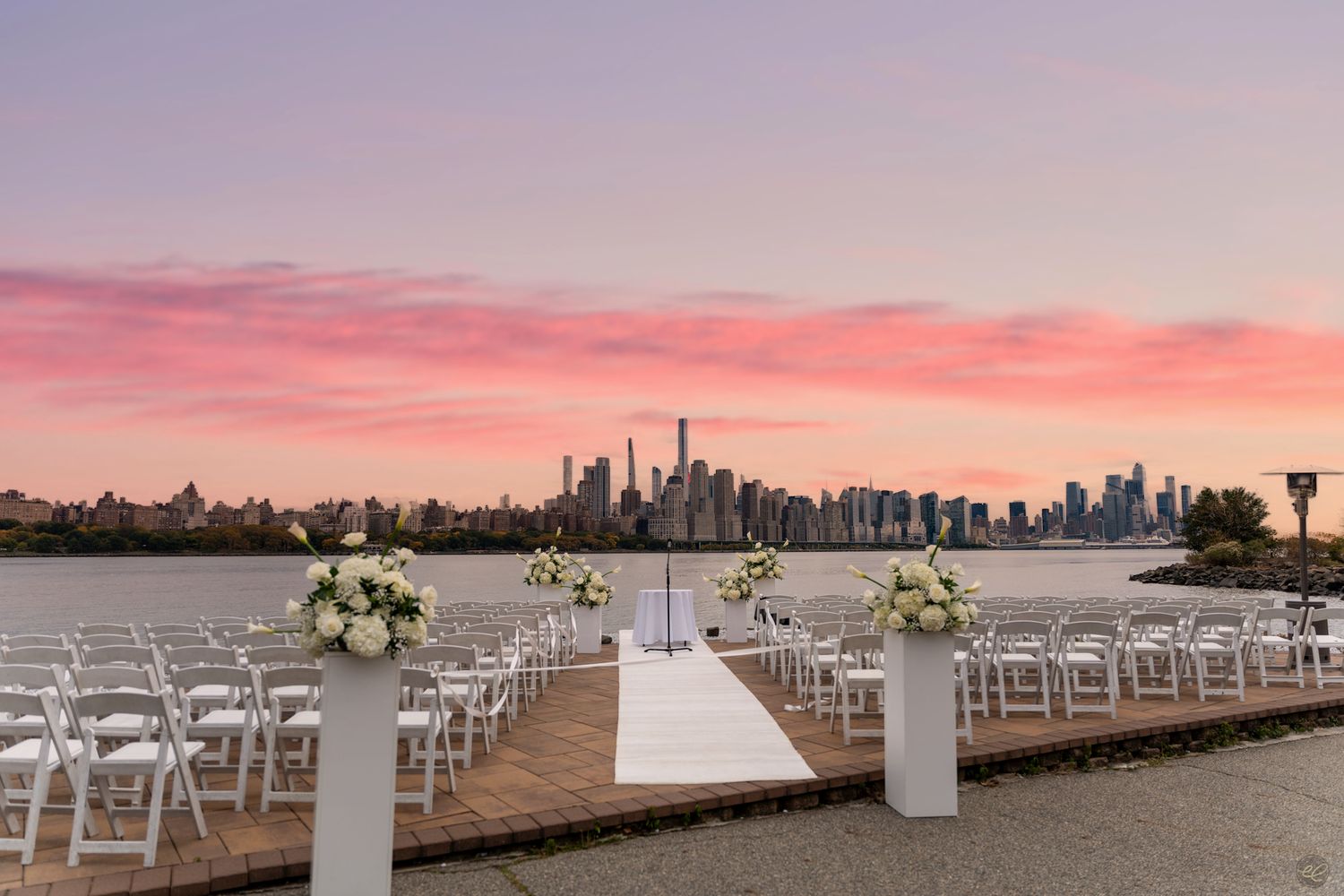 Elegant outdoor wedding ceremony setup on a waterfront dock with NYC skyline at sunset.