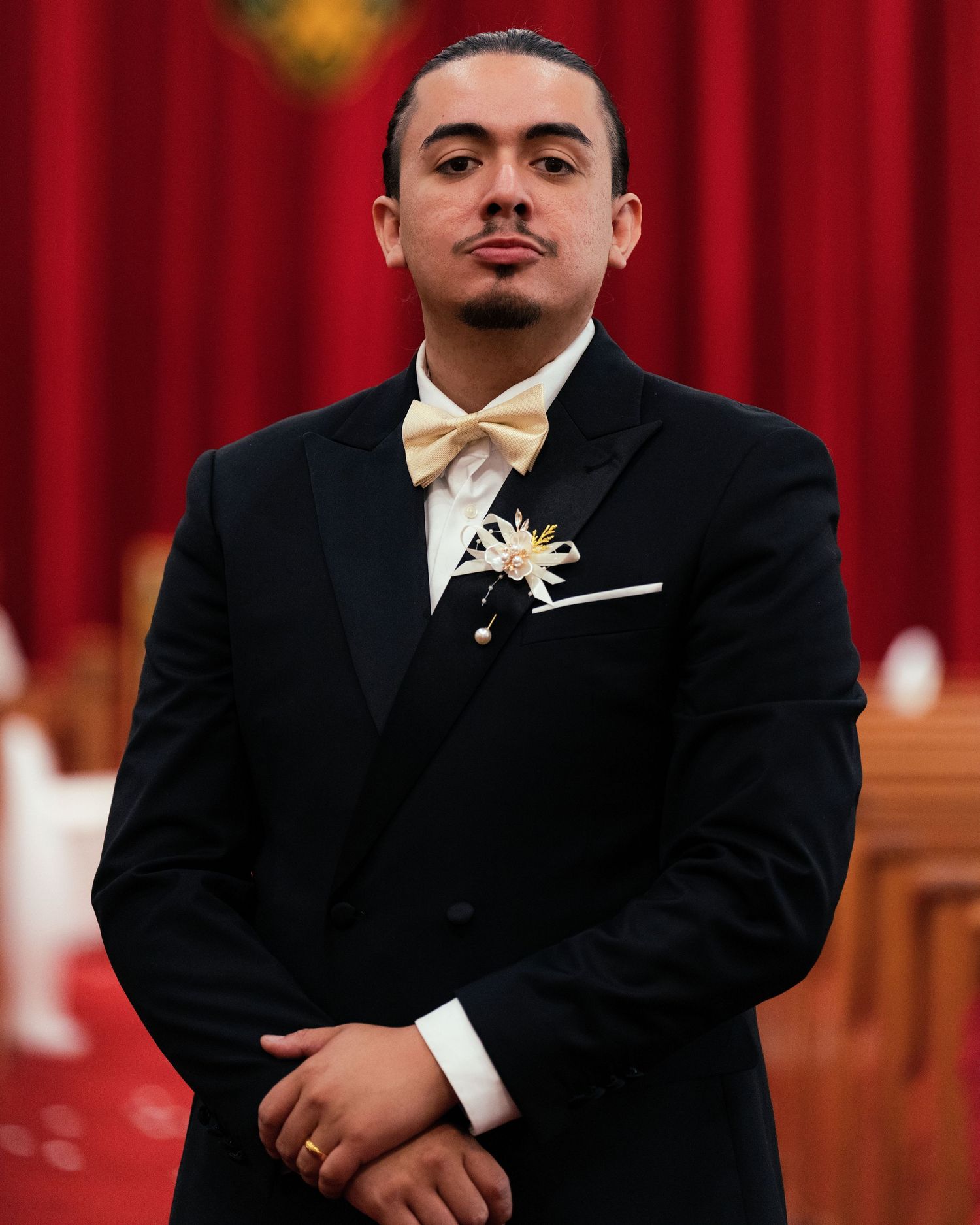 Young man in black tuxedo with cream bow tie and floral boutonniere poses formally in elegant venue.