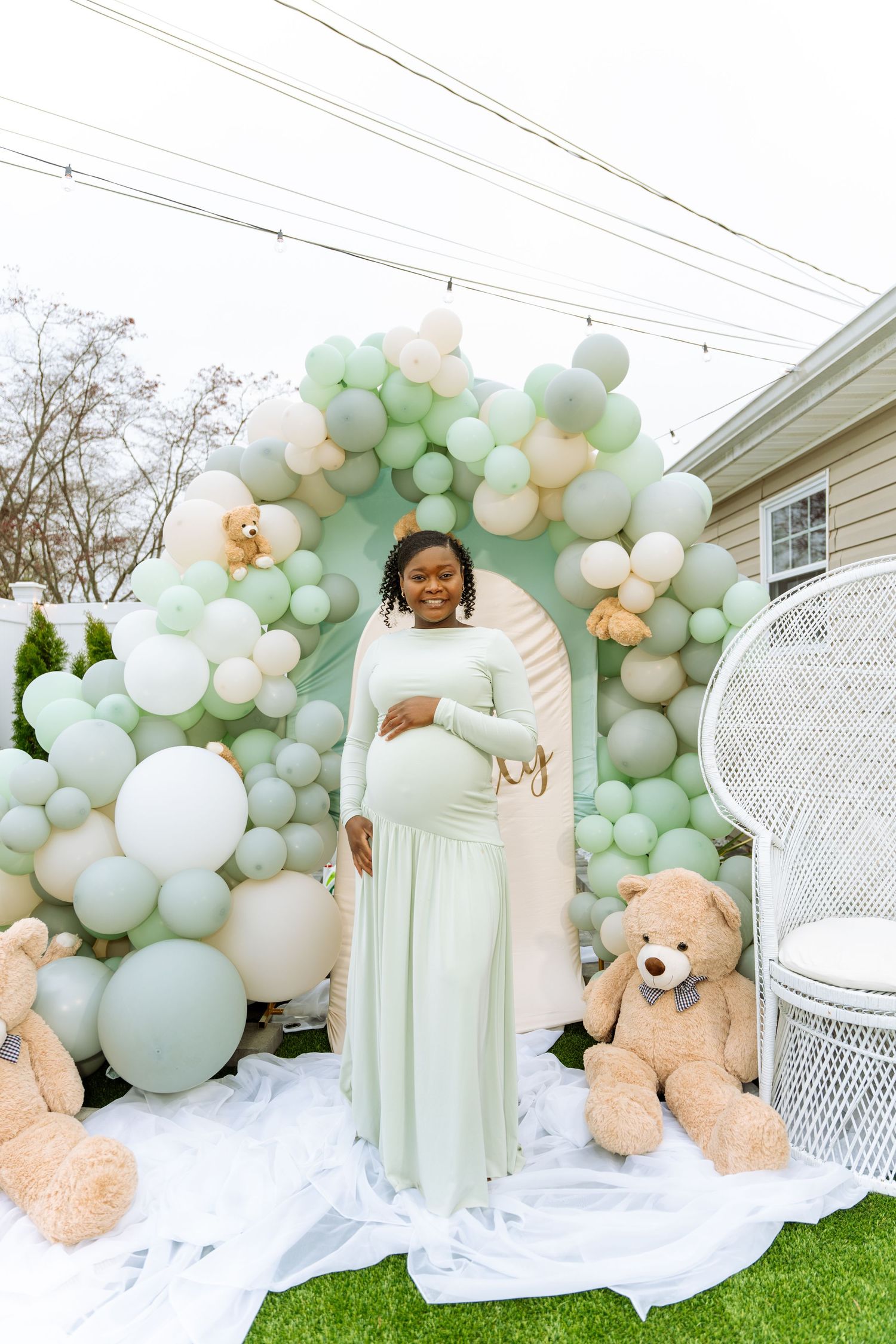 Pregnant woman in mint green gown poses before a sage balloon arch with teddy bears at a baby shower.