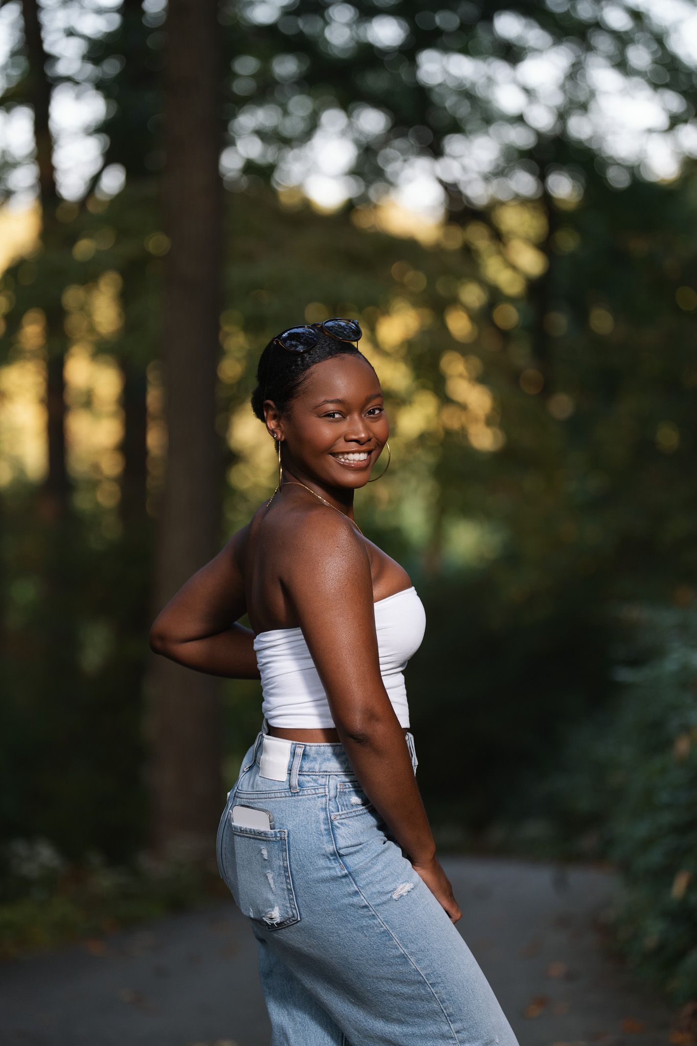 Smiling young Black woman in white tube top and jeans poses on a tree-lined path in golden evening light.