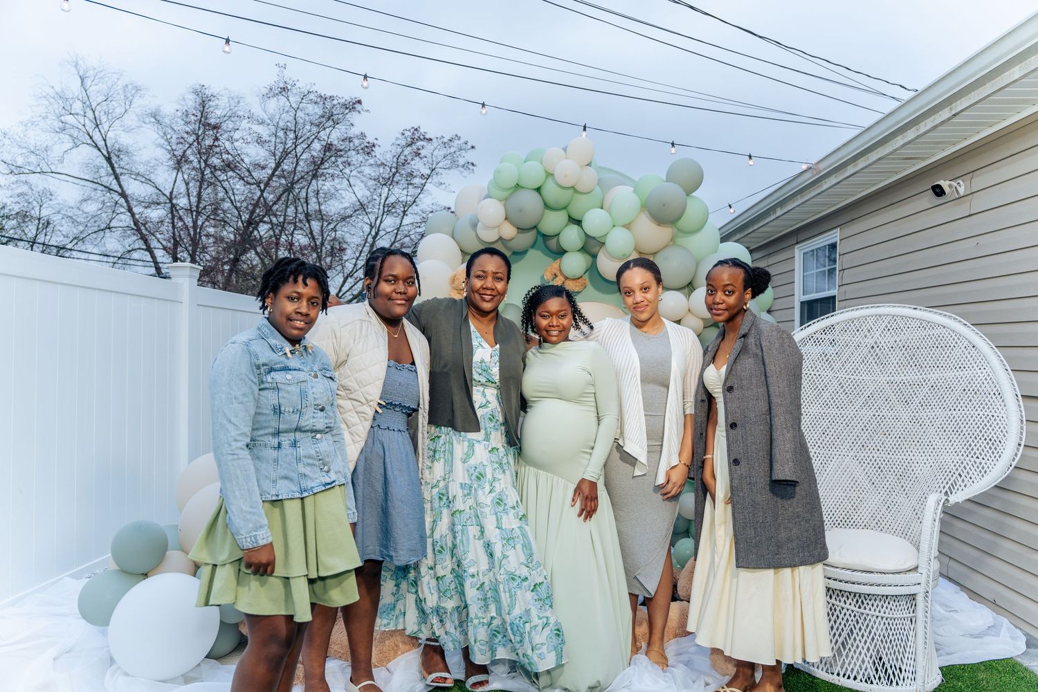 Six women pose together at an outdoor baby shower with green balloon arch backdrop and white peacock chair.