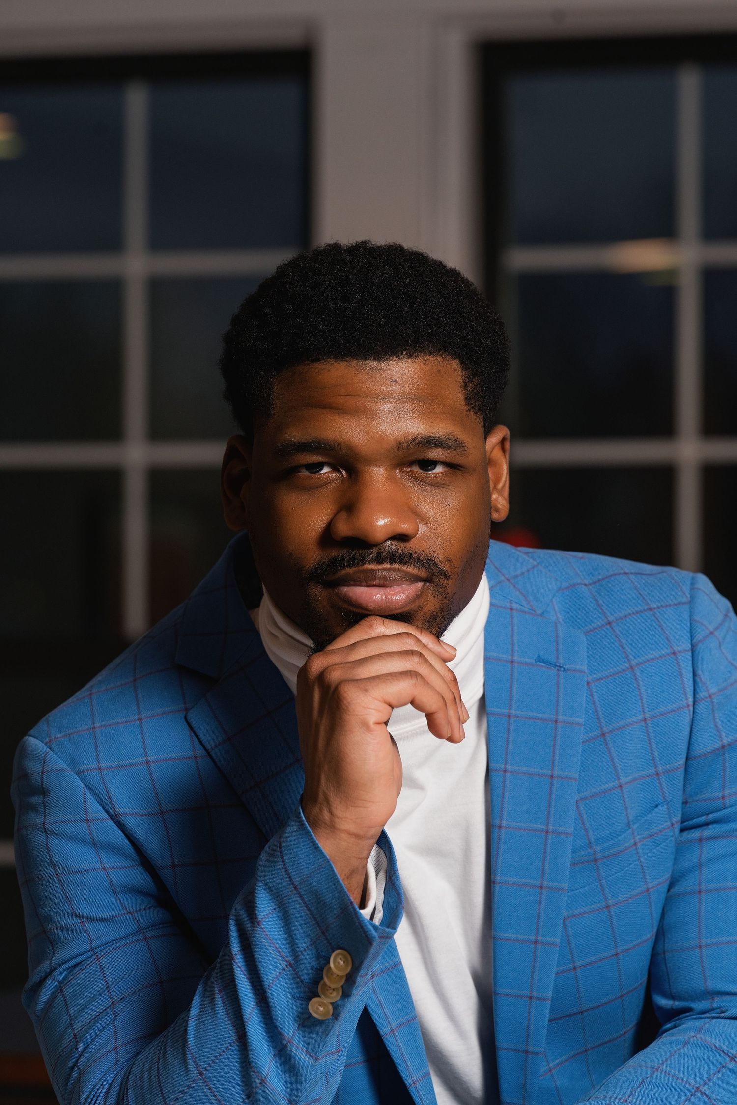 Confident Black man in blue plaid blazer poses thoughtfully with hand on chin in a professional portrait.