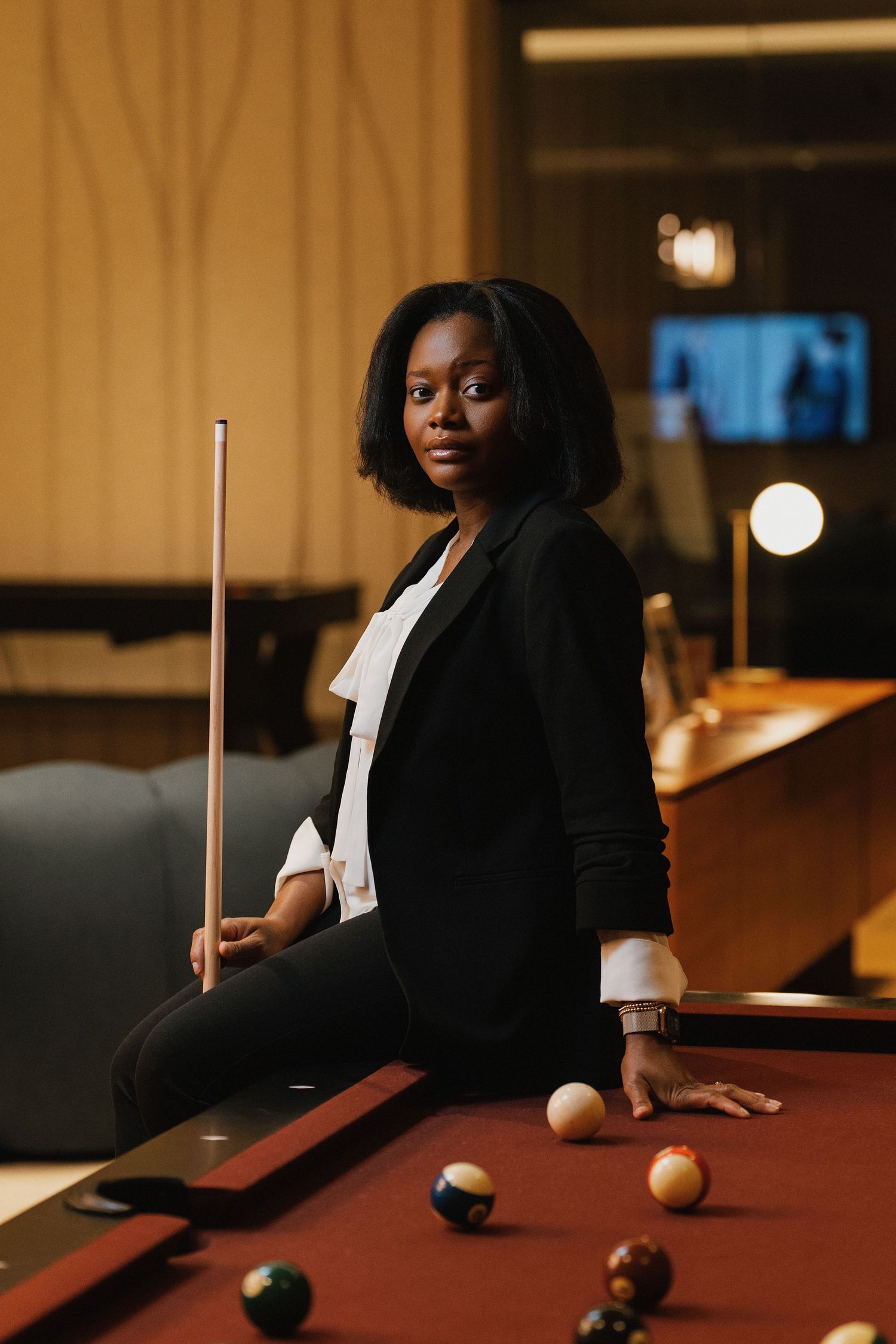 A stylish woman in a black blazer poses with a pool cue beside a billiard table with balls in a dimly lit lounge.