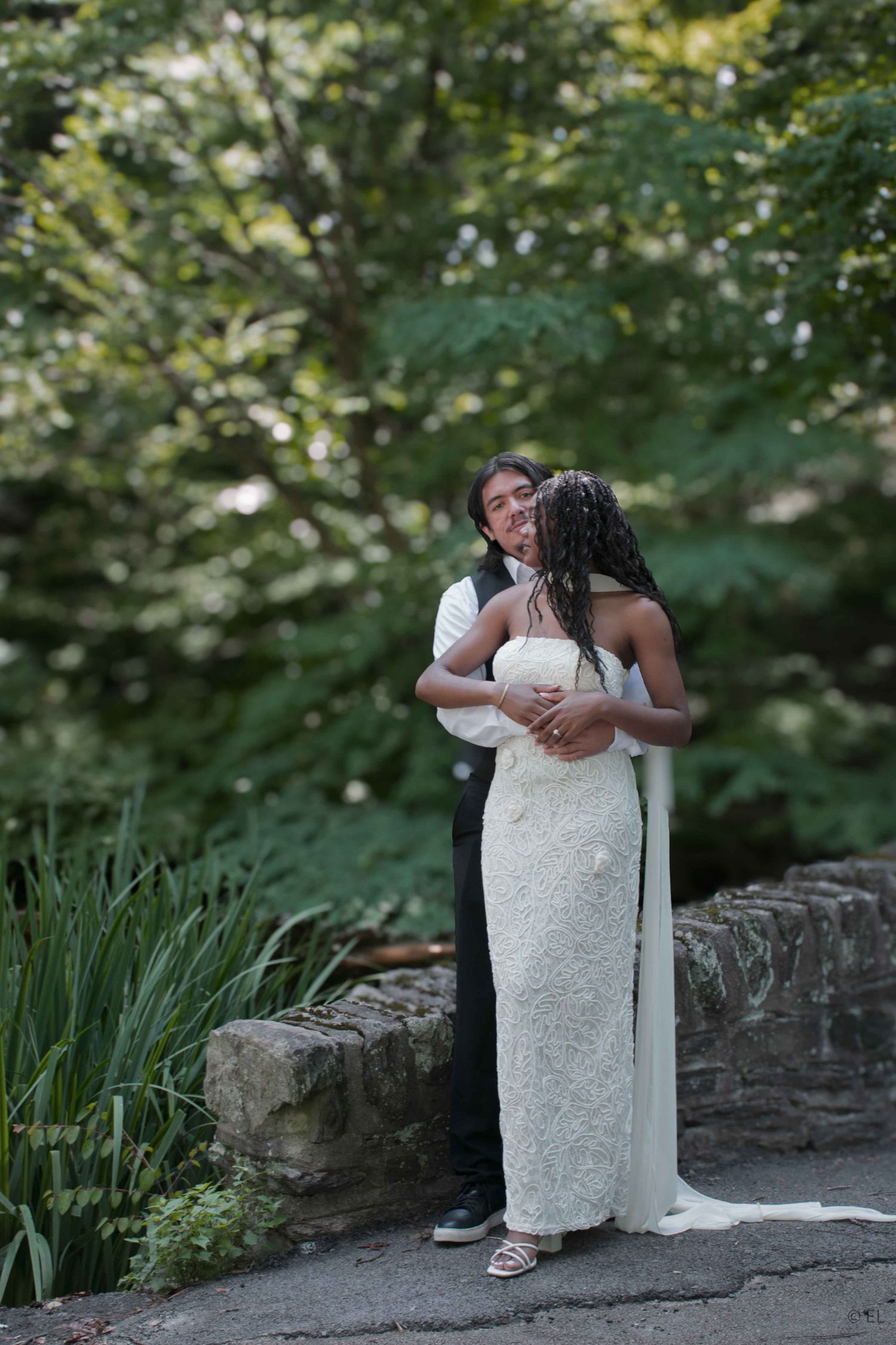 A couple in wedding attire embracing outdoors near a stone wall surrounded by lush green trees.