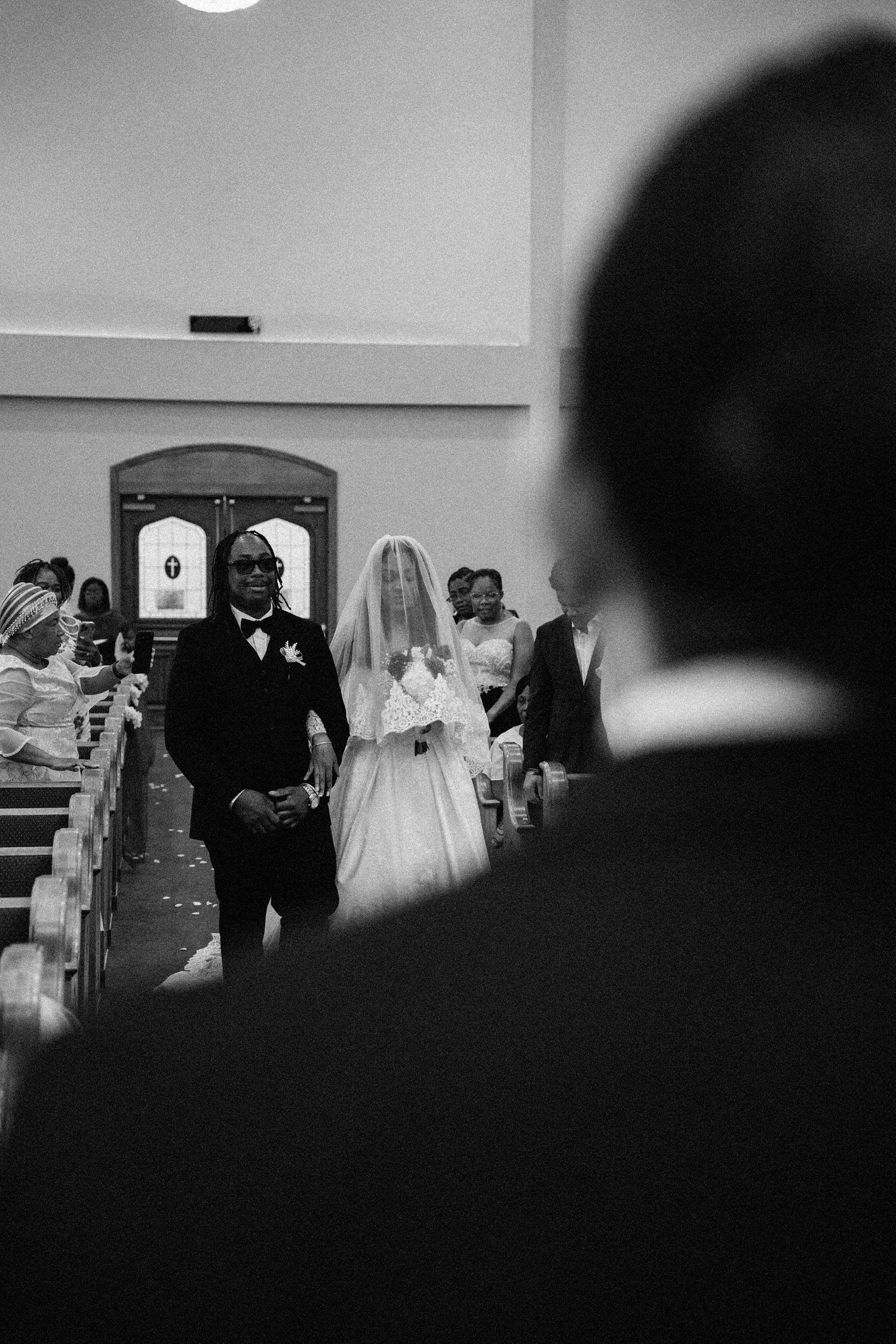 Black and white photo of a bride walking down the church aisle with escort, groom watching from foreground.