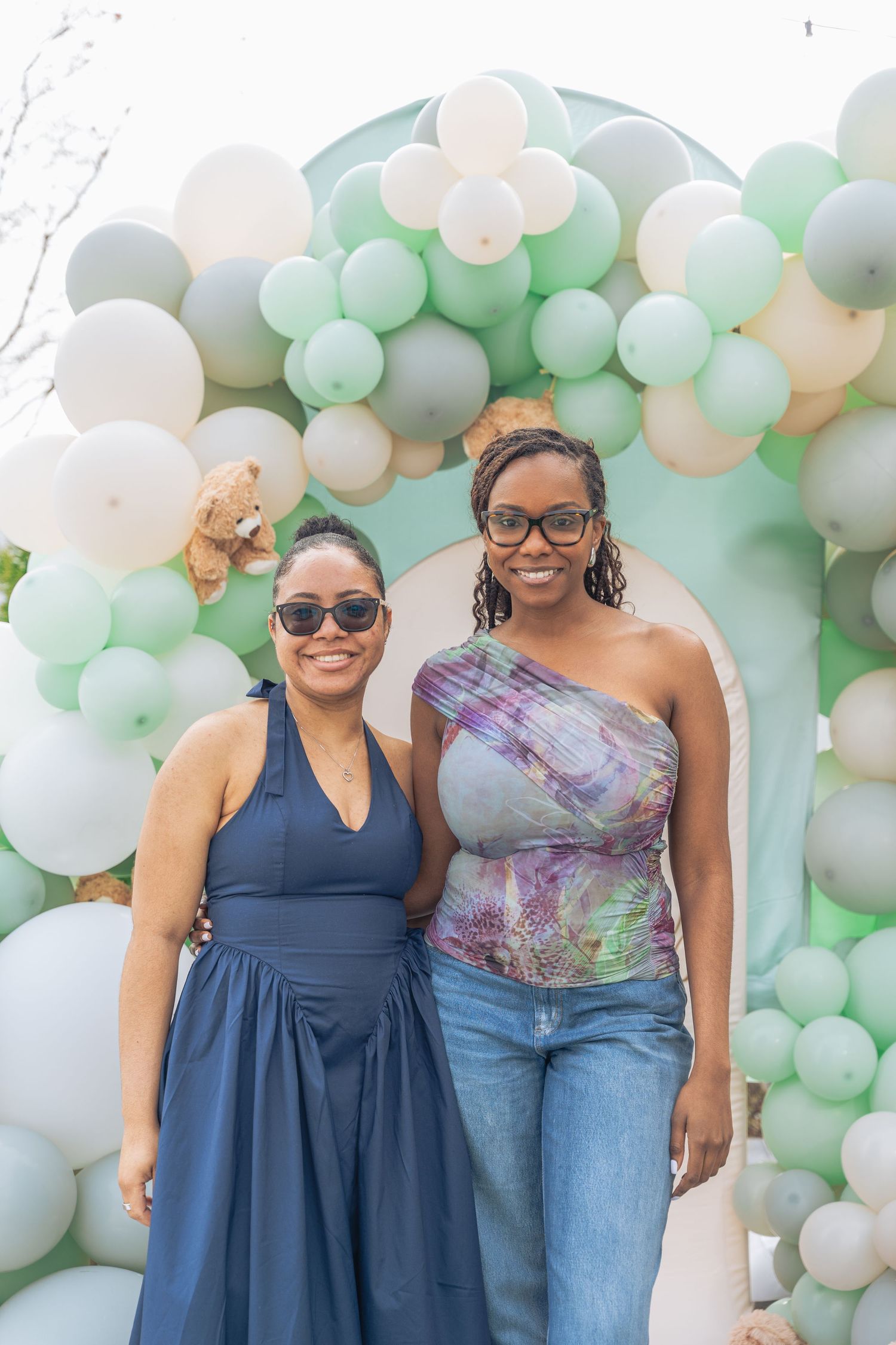 Two women smiling in front of a green and white balloon arch at an outdoor celebration.