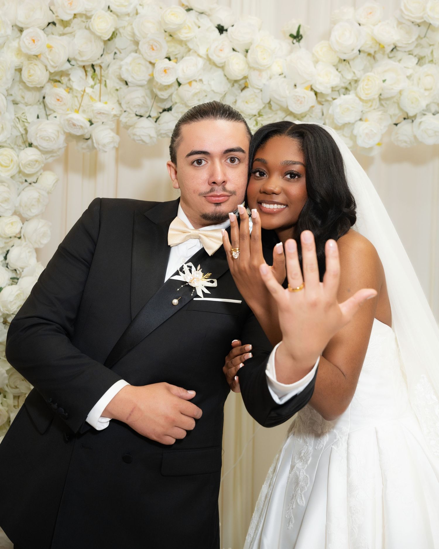 Newlywed couple in wedding attire showing off rings, standing before white floral backdrop.
