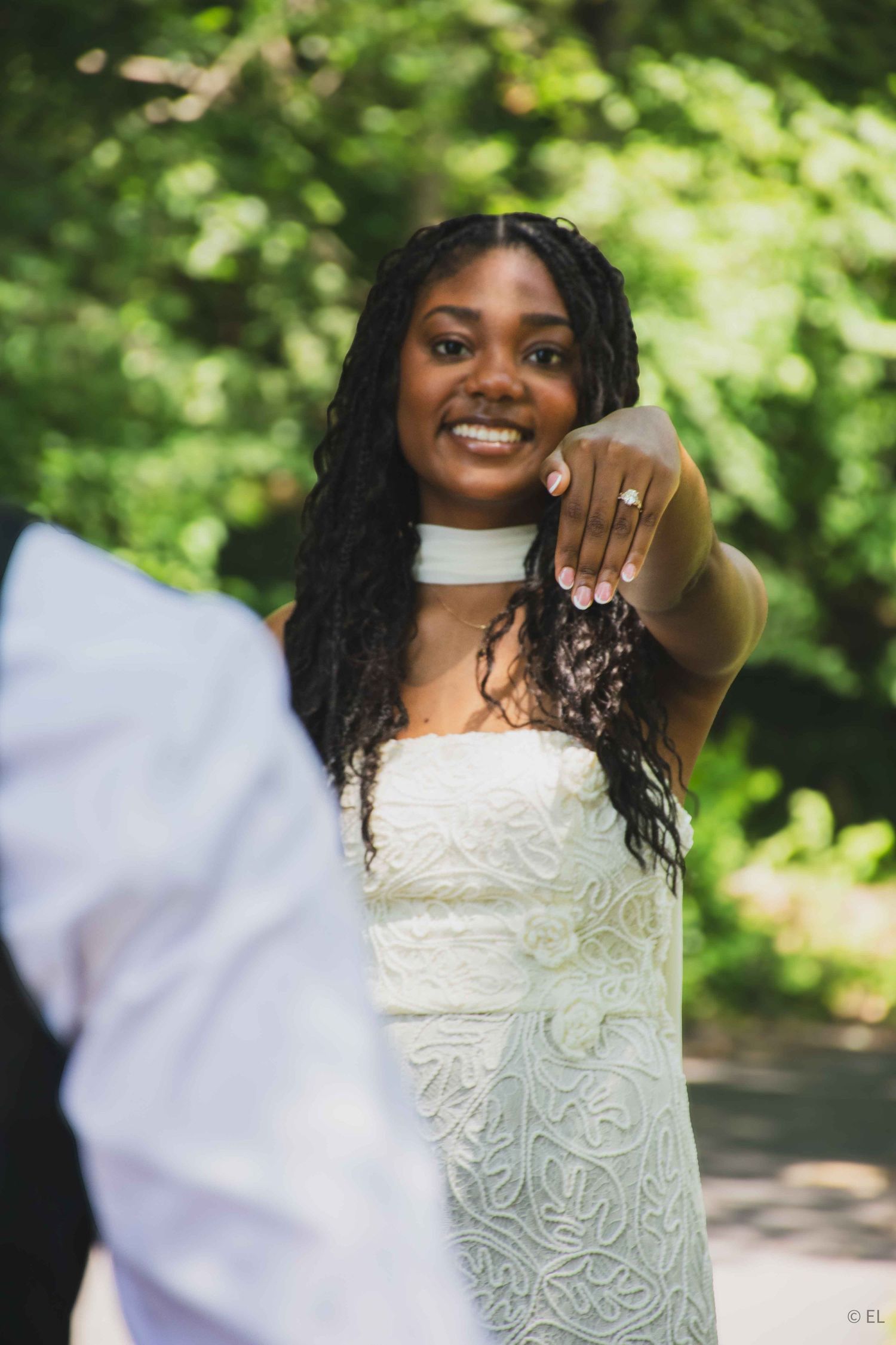 Smiling bride in white lace gown extends her hand showing wedding ring toward the camera in a green outdoor setting.