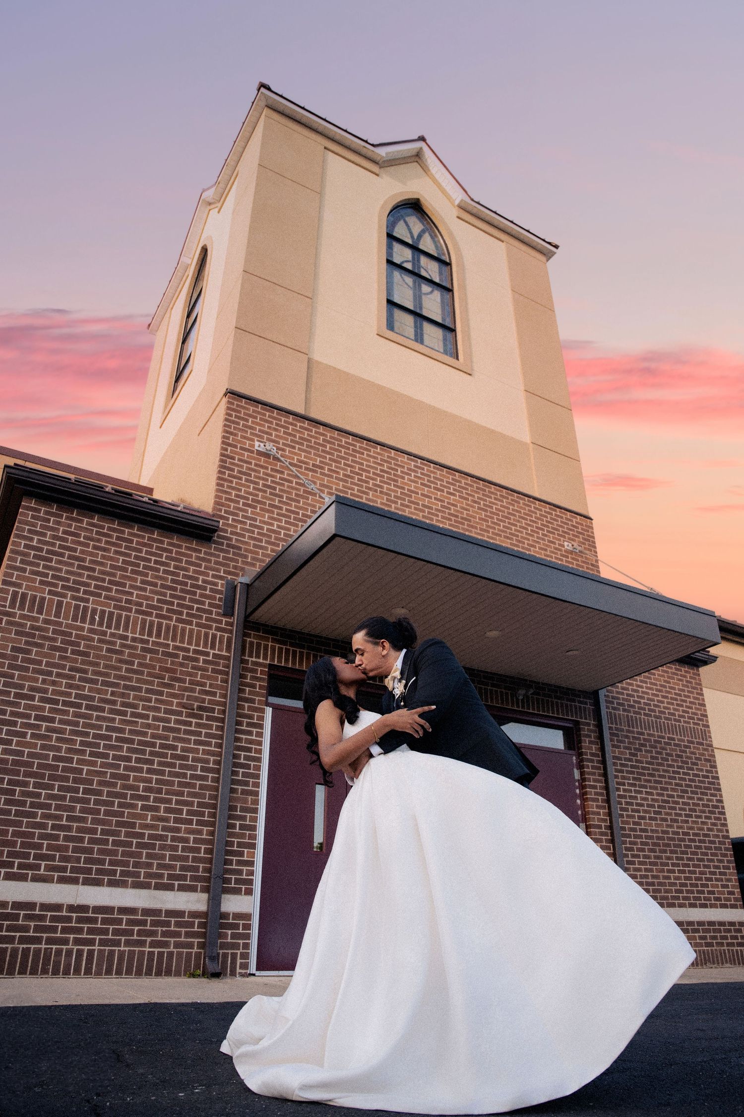 Bride and groom share a romantic dip kiss outside a brick church at sunset with a colorful pink sky backdrop.