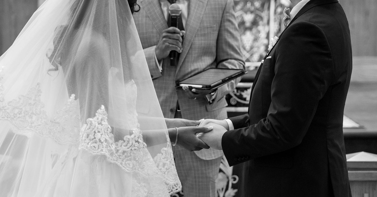 Black and white wedding ceremony photo of bride in lace veil and groom holding hands before officiant.