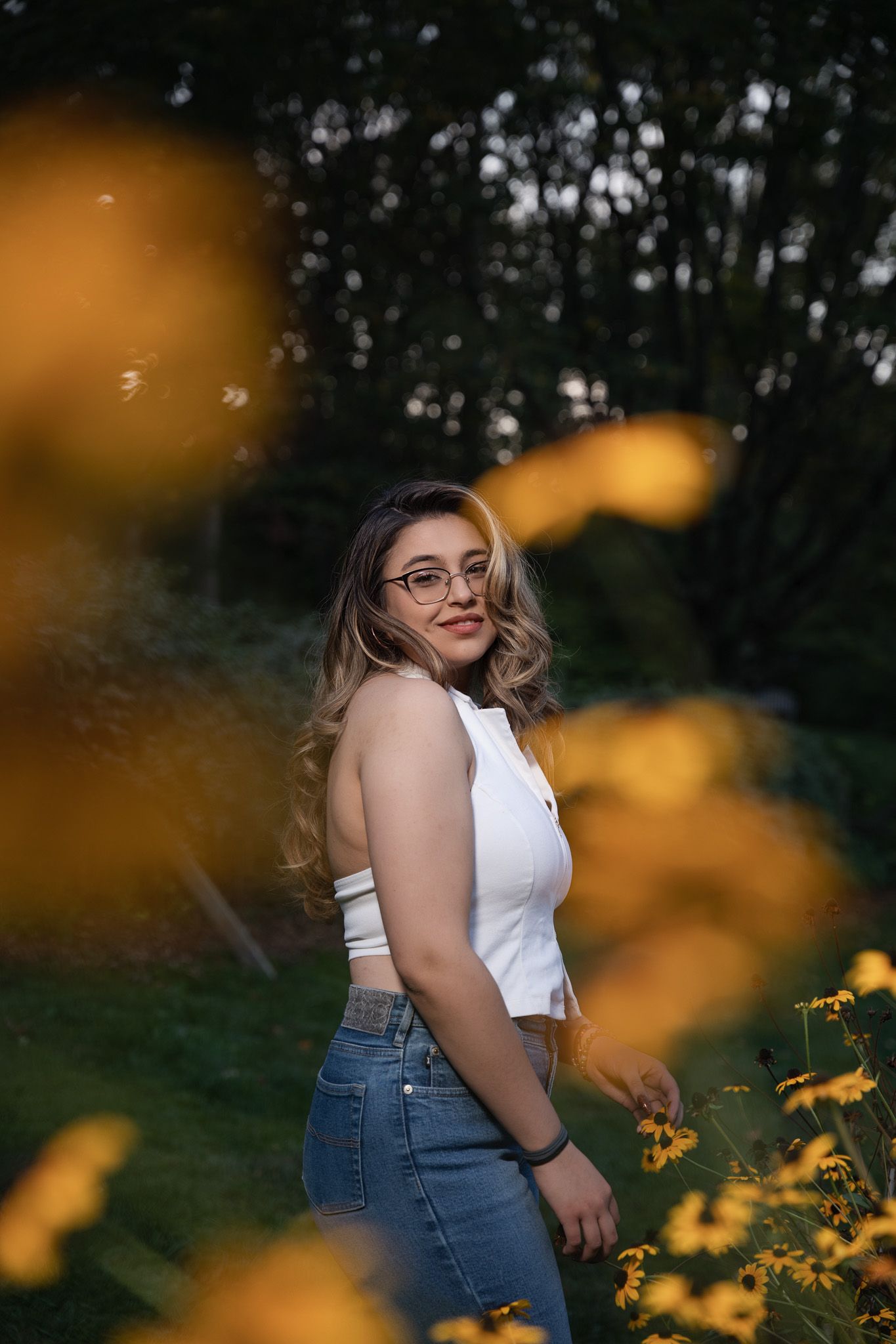 Young woman with glasses and wavy blonde hair poses among yellow flowers in a garden wearing a white crop top and jeans.