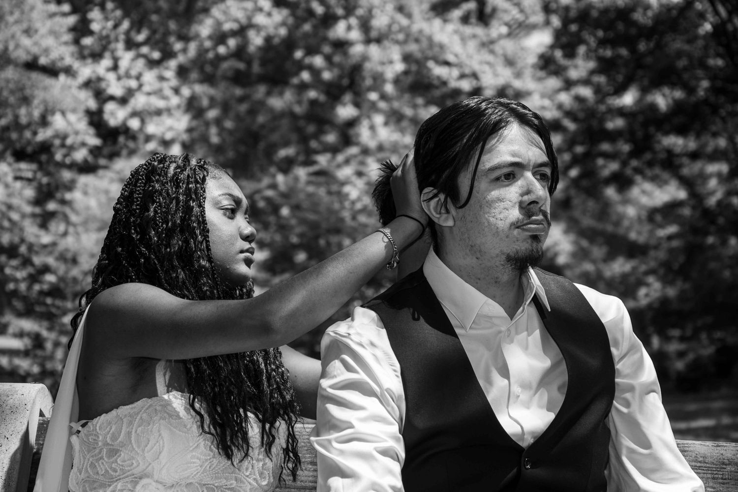 Black and white photo of a bride adjusting the hair of a groom outdoors under leafy trees.