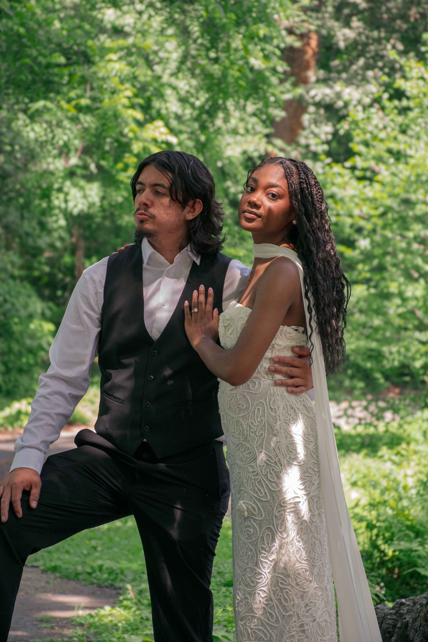 Interracial wedding couple posing together in a lush green park, bride in white lace gown and groom in black vest.