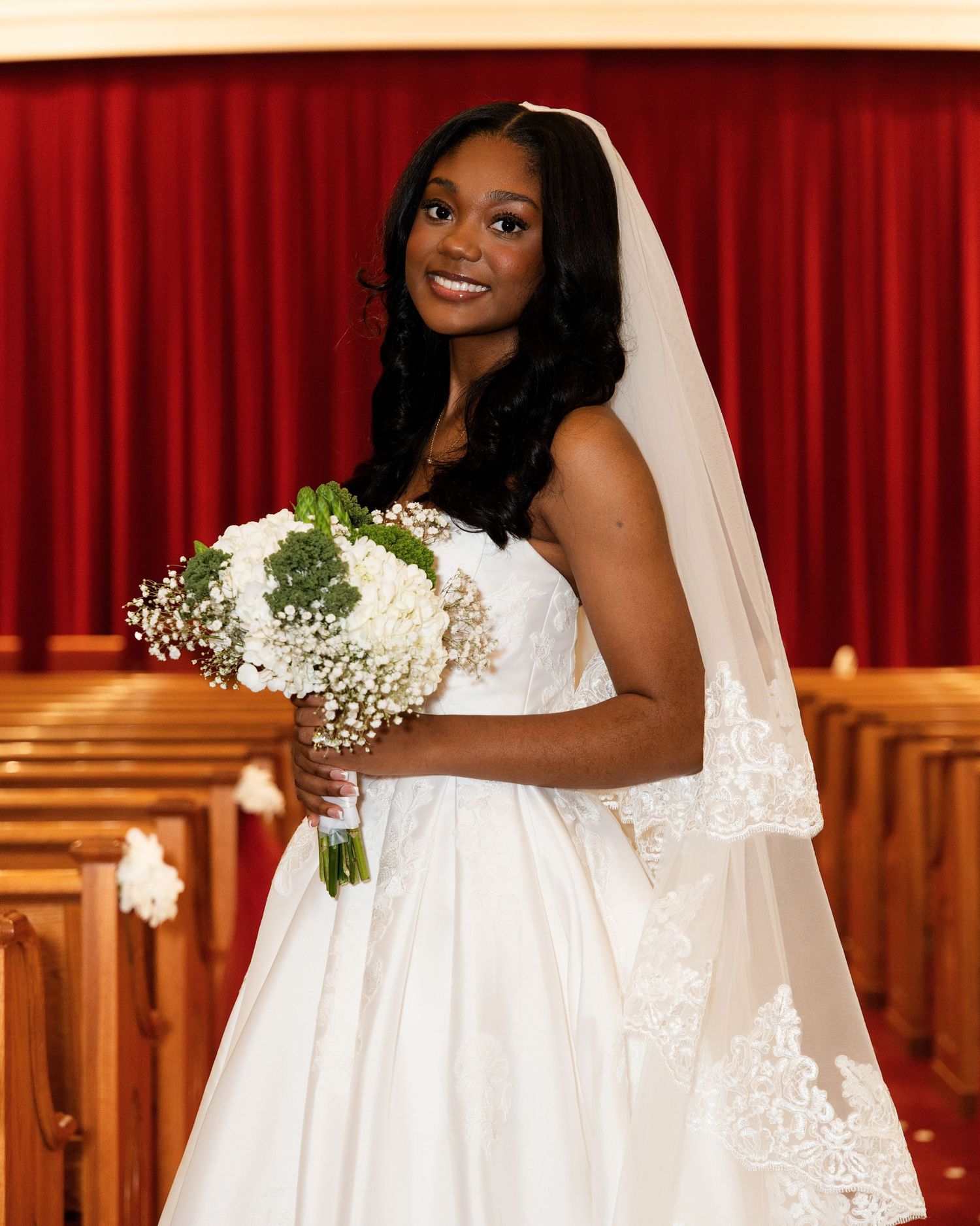 A smiling bride in a white gown and veil holds a white floral bouquet inside a church with red curtains.