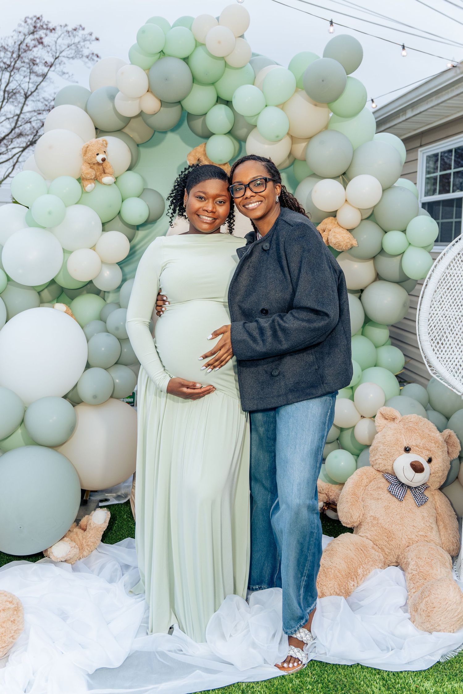 Two women pose at a baby shower with sage green balloon arch and teddy bears.