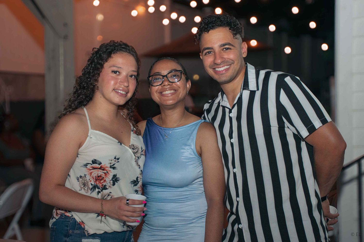 Three young friends smiling together at an outdoor evening party with string lights.