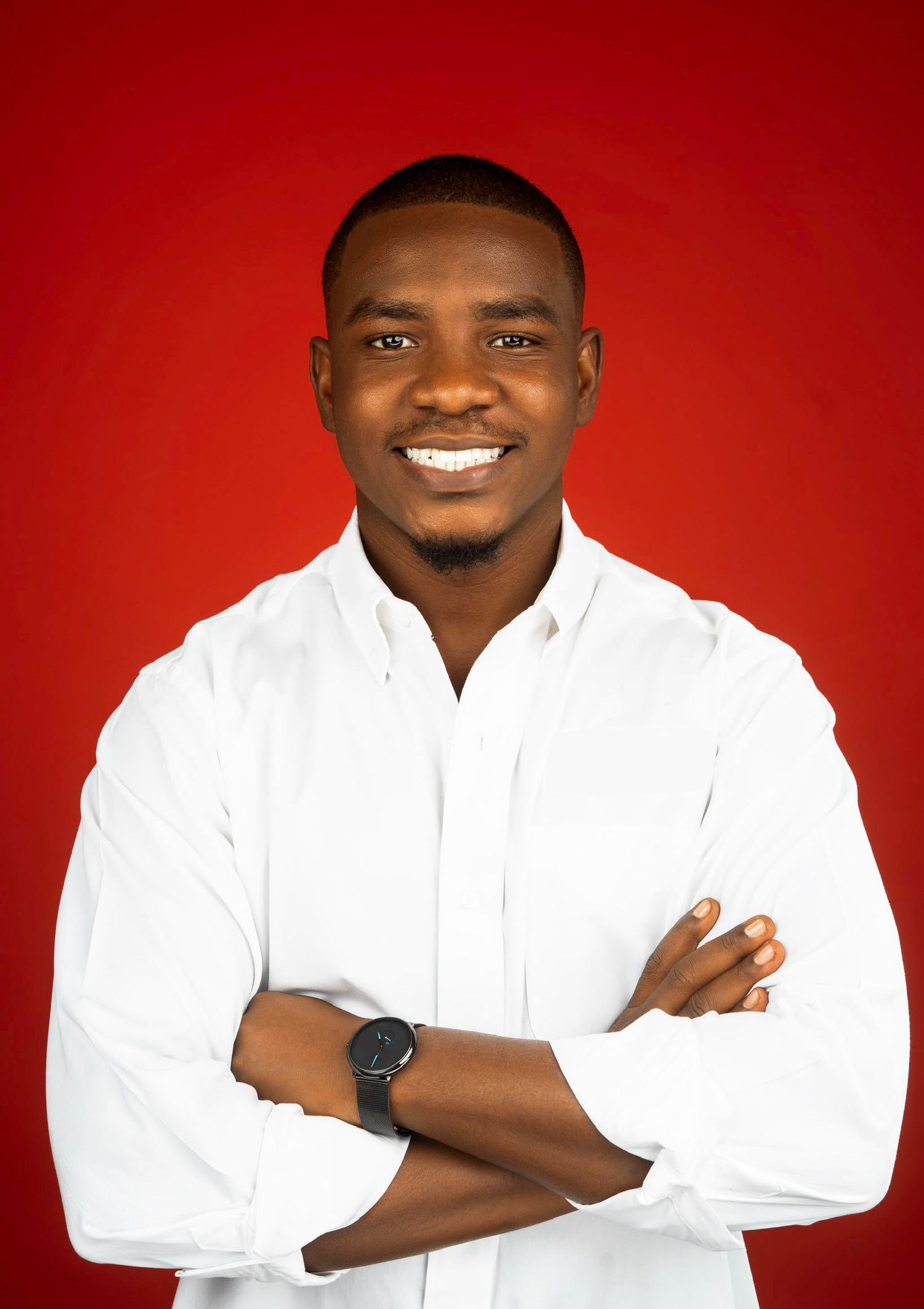 A confident young man in a white shirt with arms crossed smiling against a bold red background.