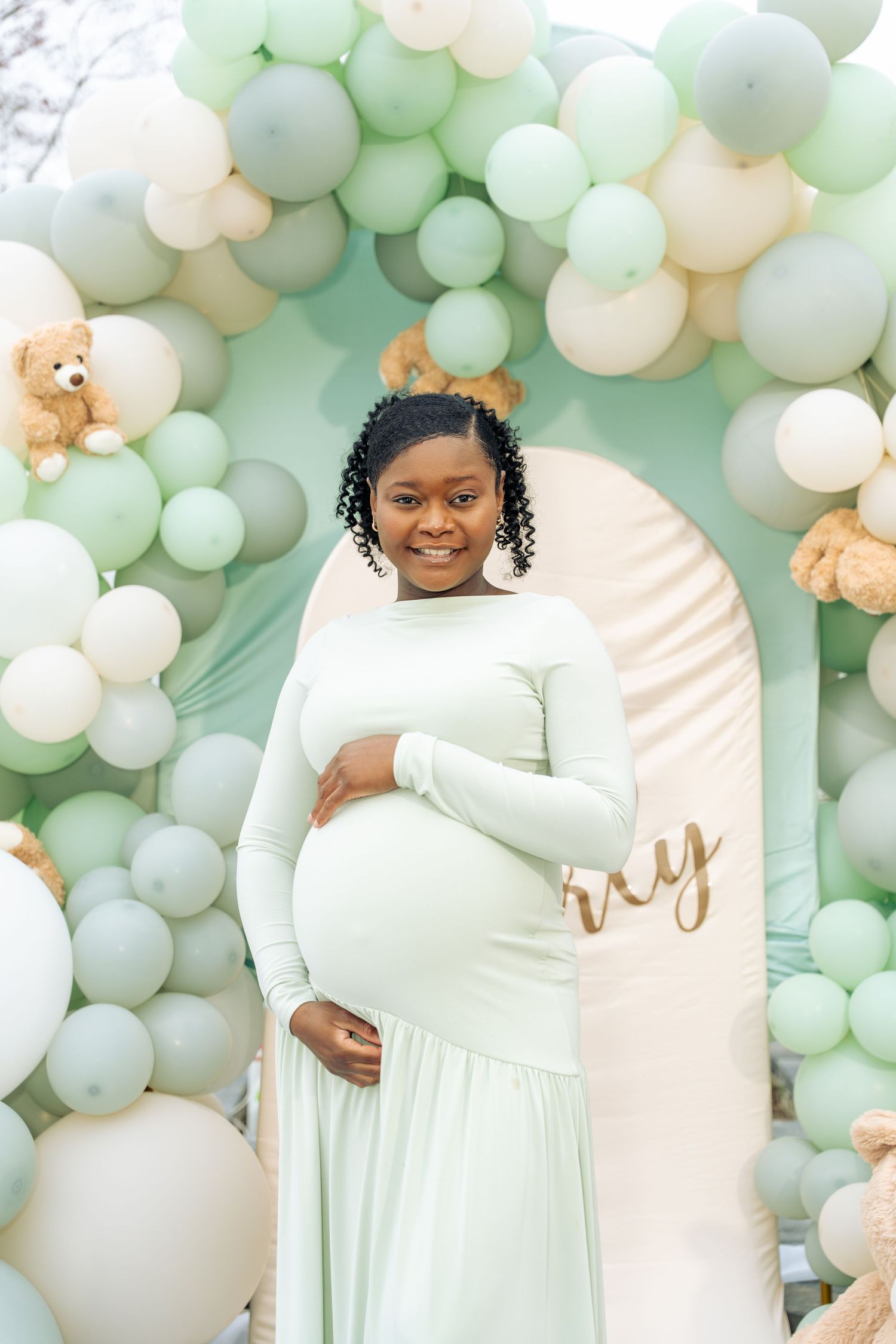Smiling pregnant woman in mint green dress at a balloon-decorated baby shower celebration.