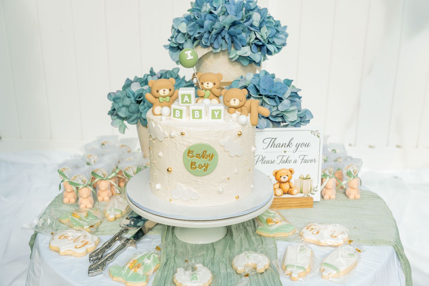 Baby boy shower cake with blue hydrangeas, teddy bears, cookies, and favor boxes on dessert table.