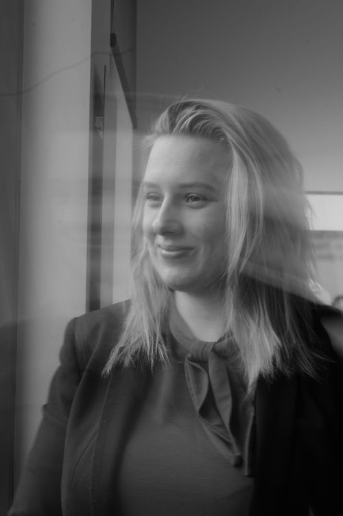 Black and white portrait of a smiling blonde woman standing by a window with soft motion blur effect.