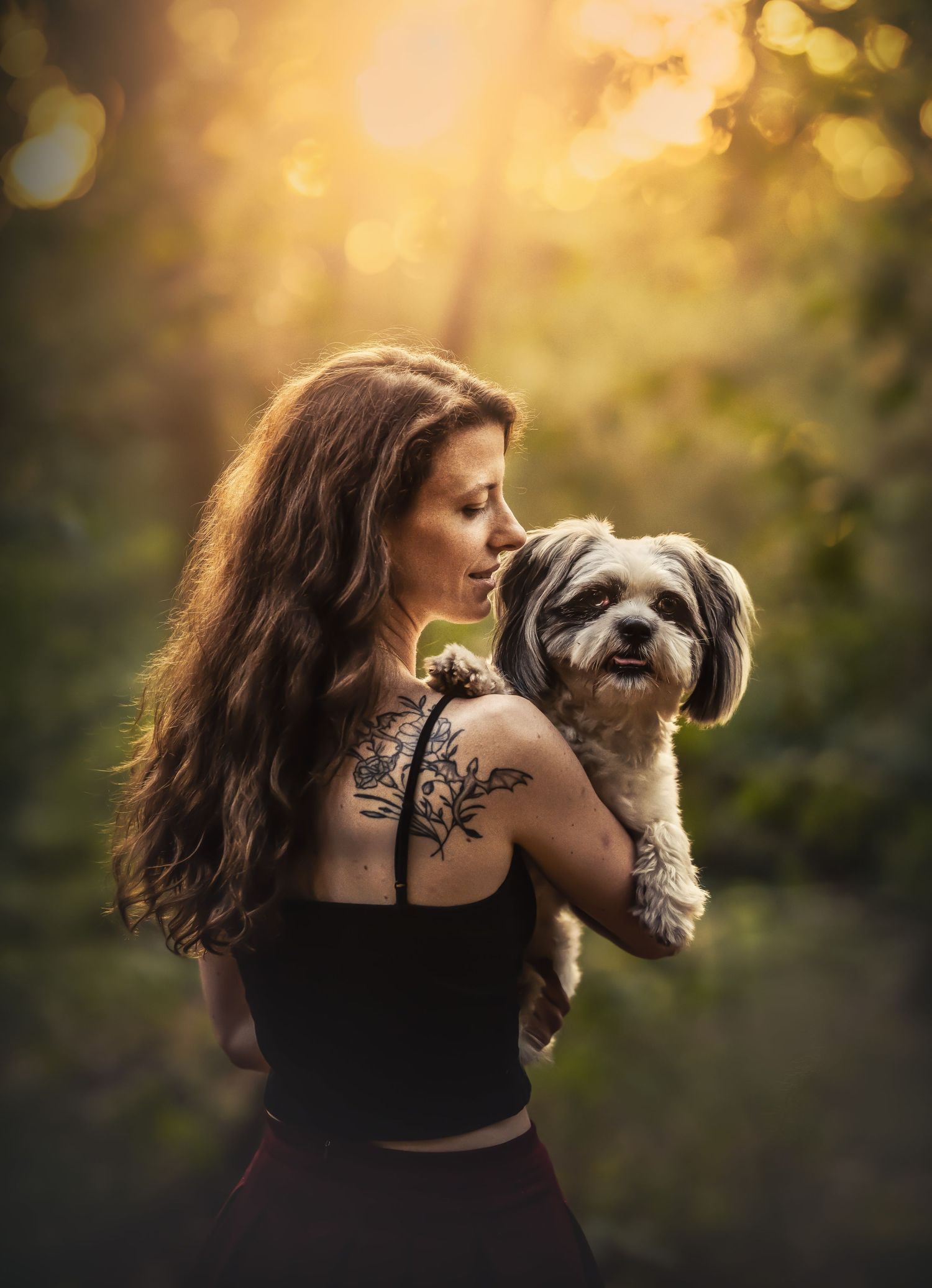 Woman with tattoos holds a small Shih Tzu dog in a sunlit forest, golden light glowing through green trees.