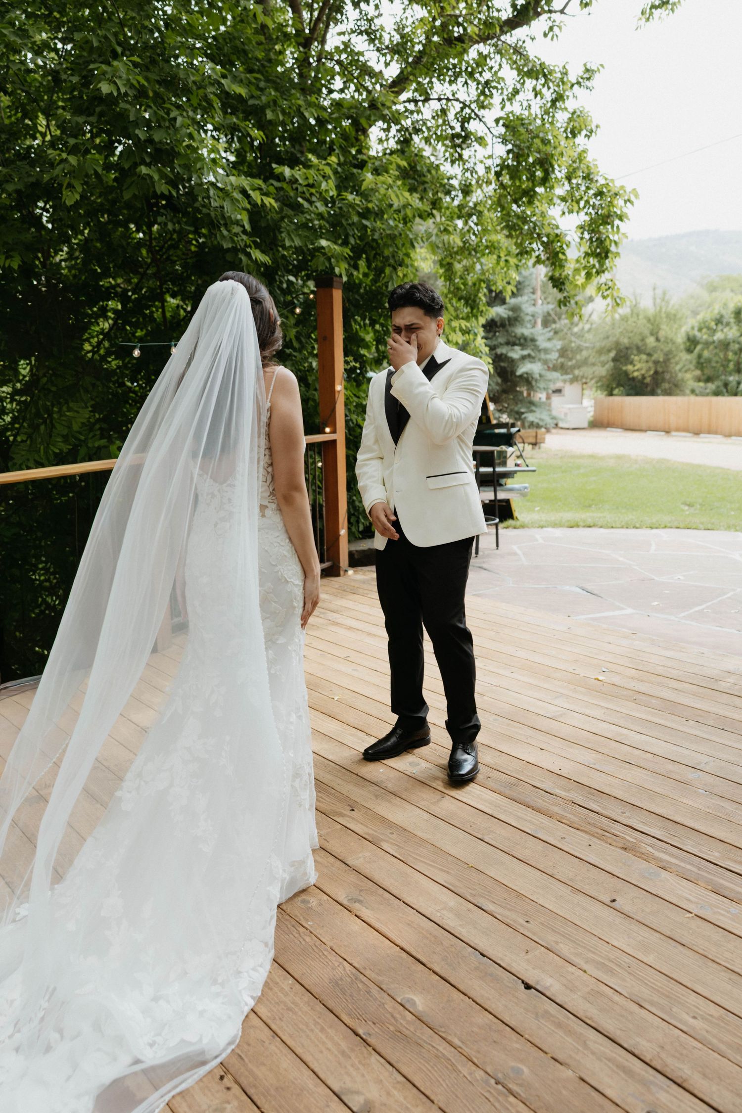 Groom in white tuxedo jacket emotionally reacts seeing bride in white gown and veil during first look on wooden deck.
