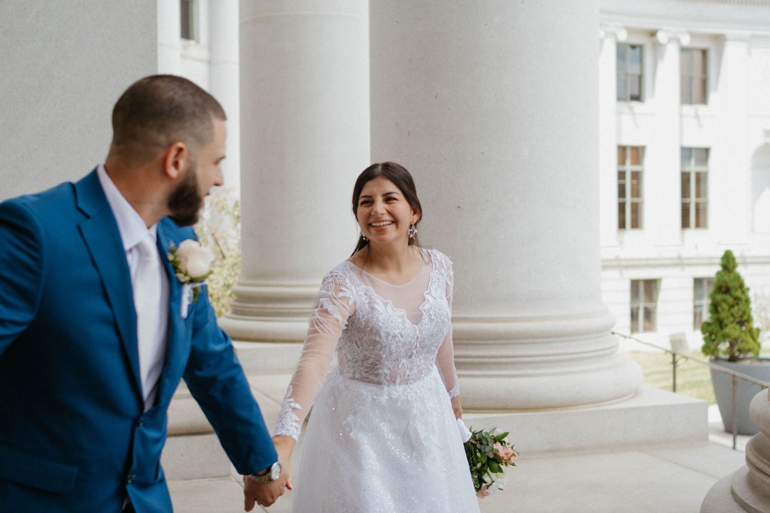 Bride in lace gown and groom in blue suit hold hands smiling at outdoor wedding venue with white columns.