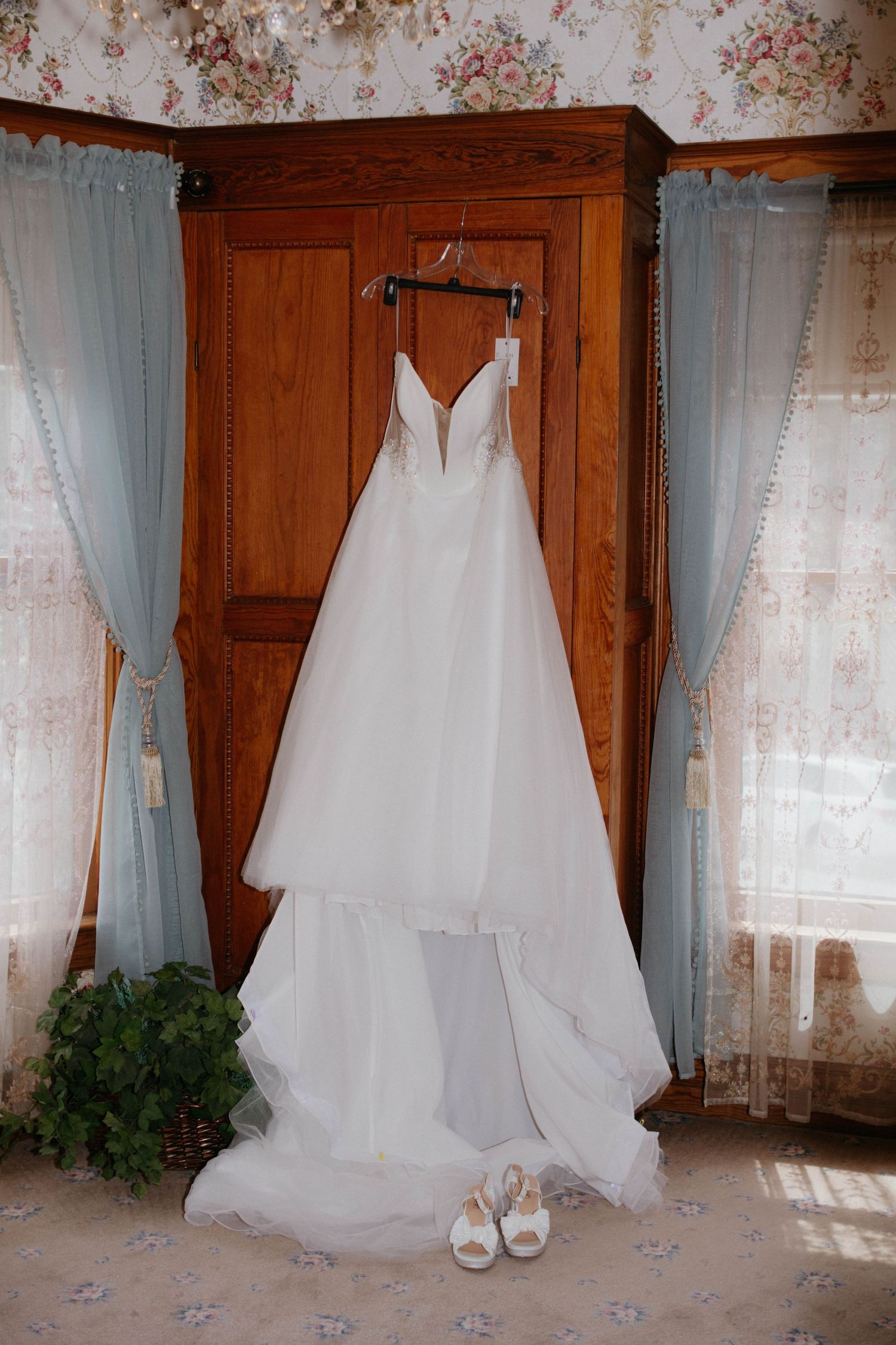A white wedding dress with plunging neckline hangs on wooden wardrobe with matching shoes beneath and sheer curtains.
