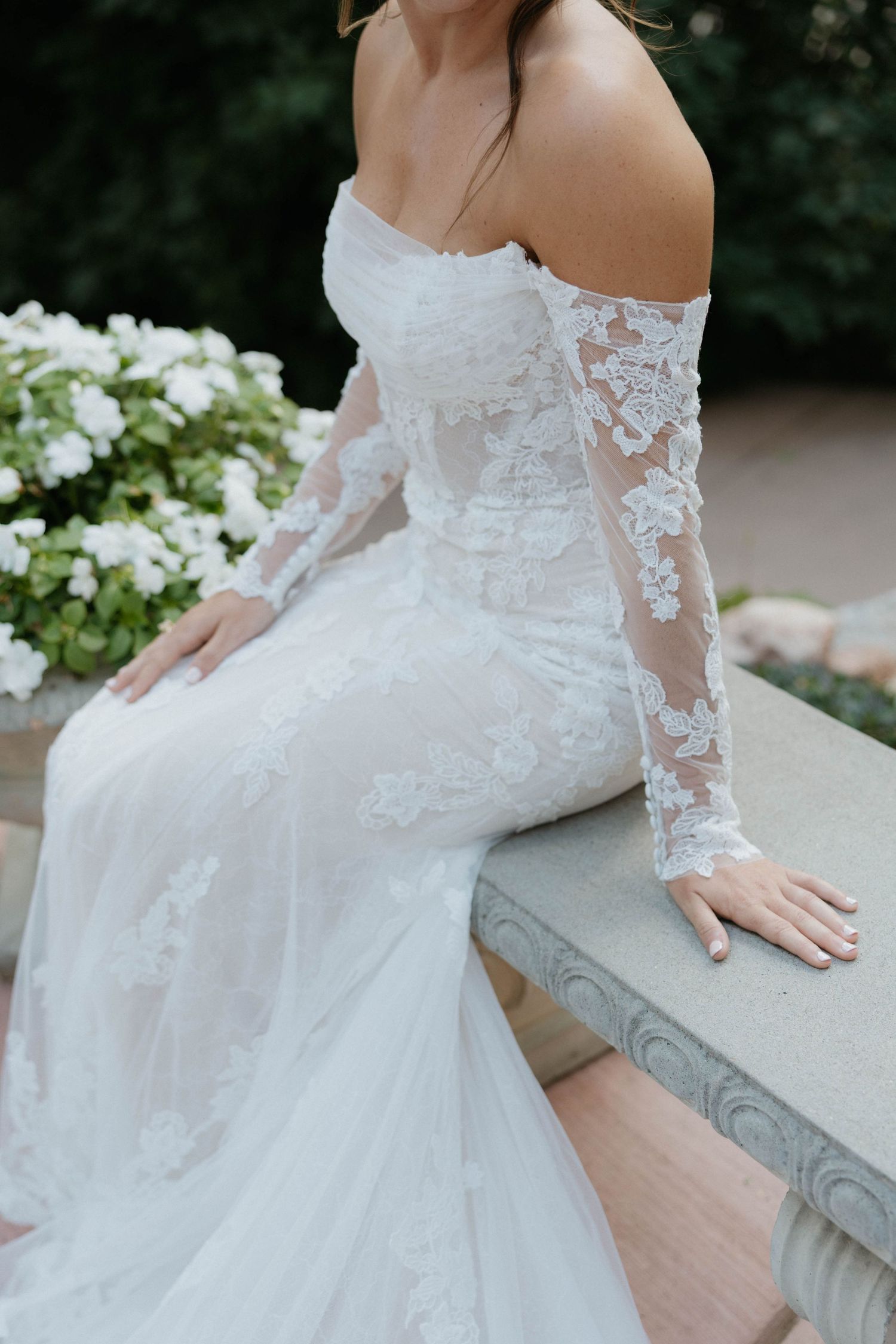 Bride in off-shoulder lace wedding gown with sheer sleeves sits on stone bench surrounded by white flowers.