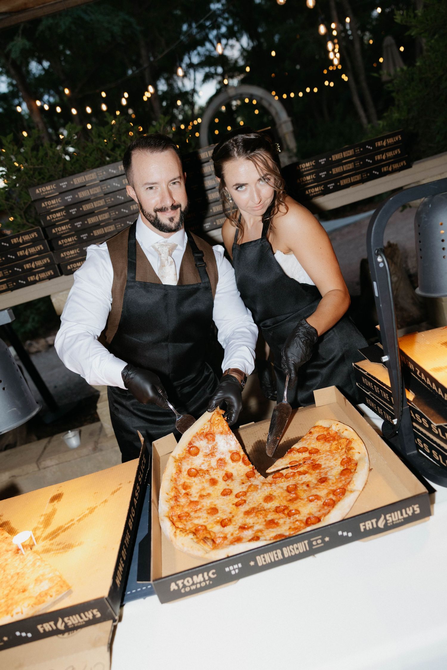 Couple in black aprons serving heart-shaped pepperoni pizza from Atomic Pizza box at outdoor wedding reception.