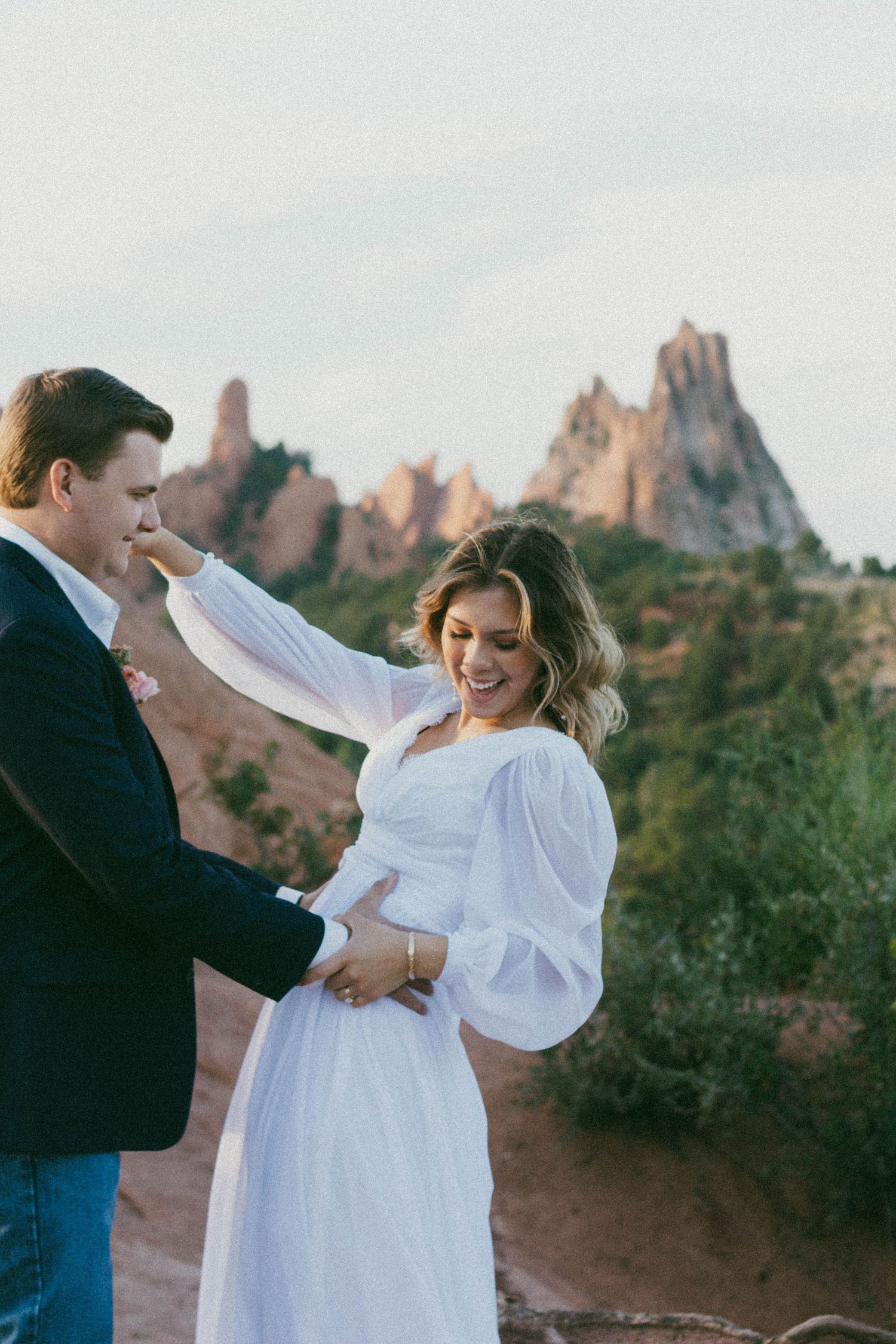 Couple sharing a romantic moment at Garden of the Gods, woman in white dress laughing joyfully with red rock formations behind.
