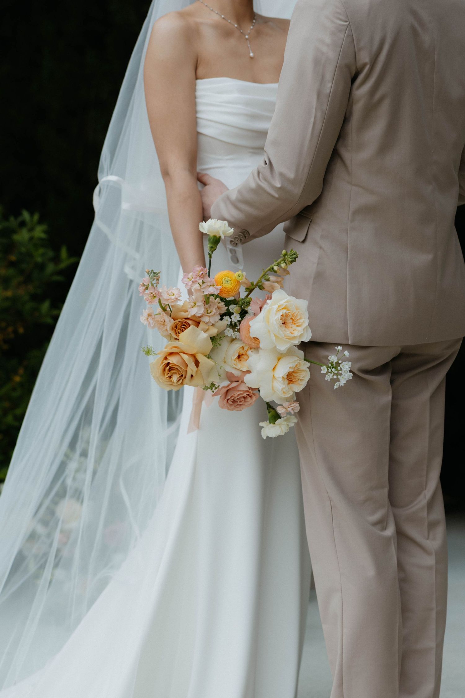 Bride in white strapless gown with veil holds peach and yellow floral bouquet beside groom in beige suit.