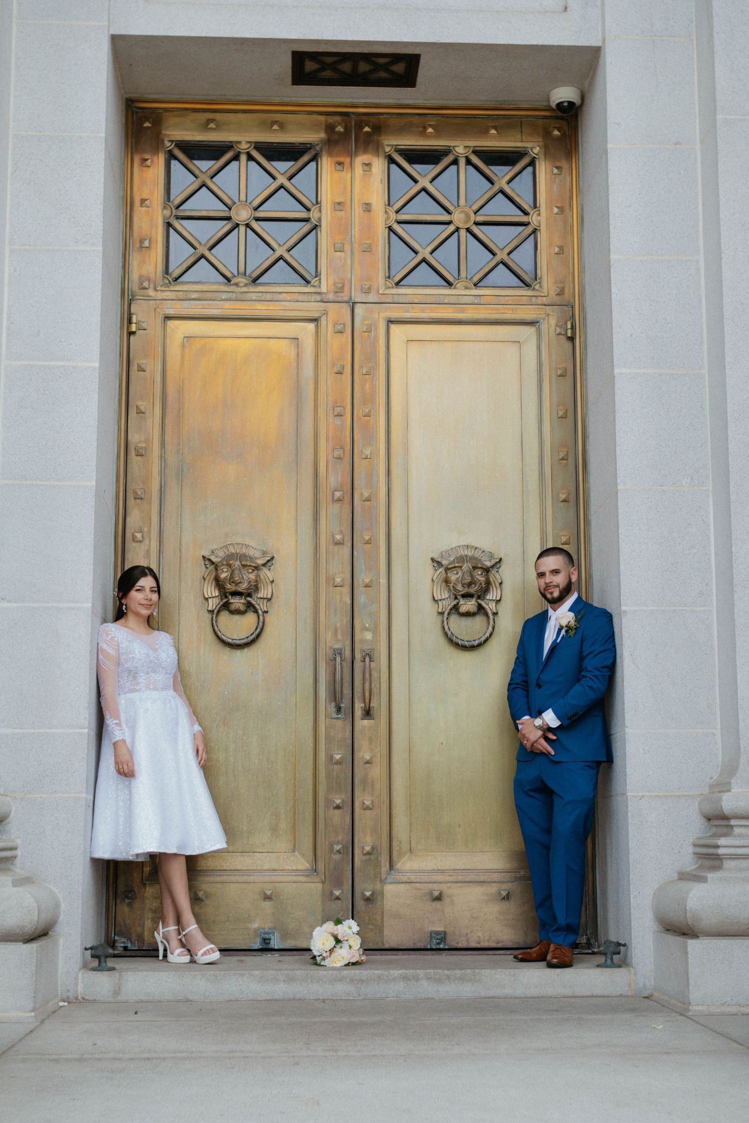 Bride and groom posing on either side of ornate golden doors with lion knockers at a classic stone building.