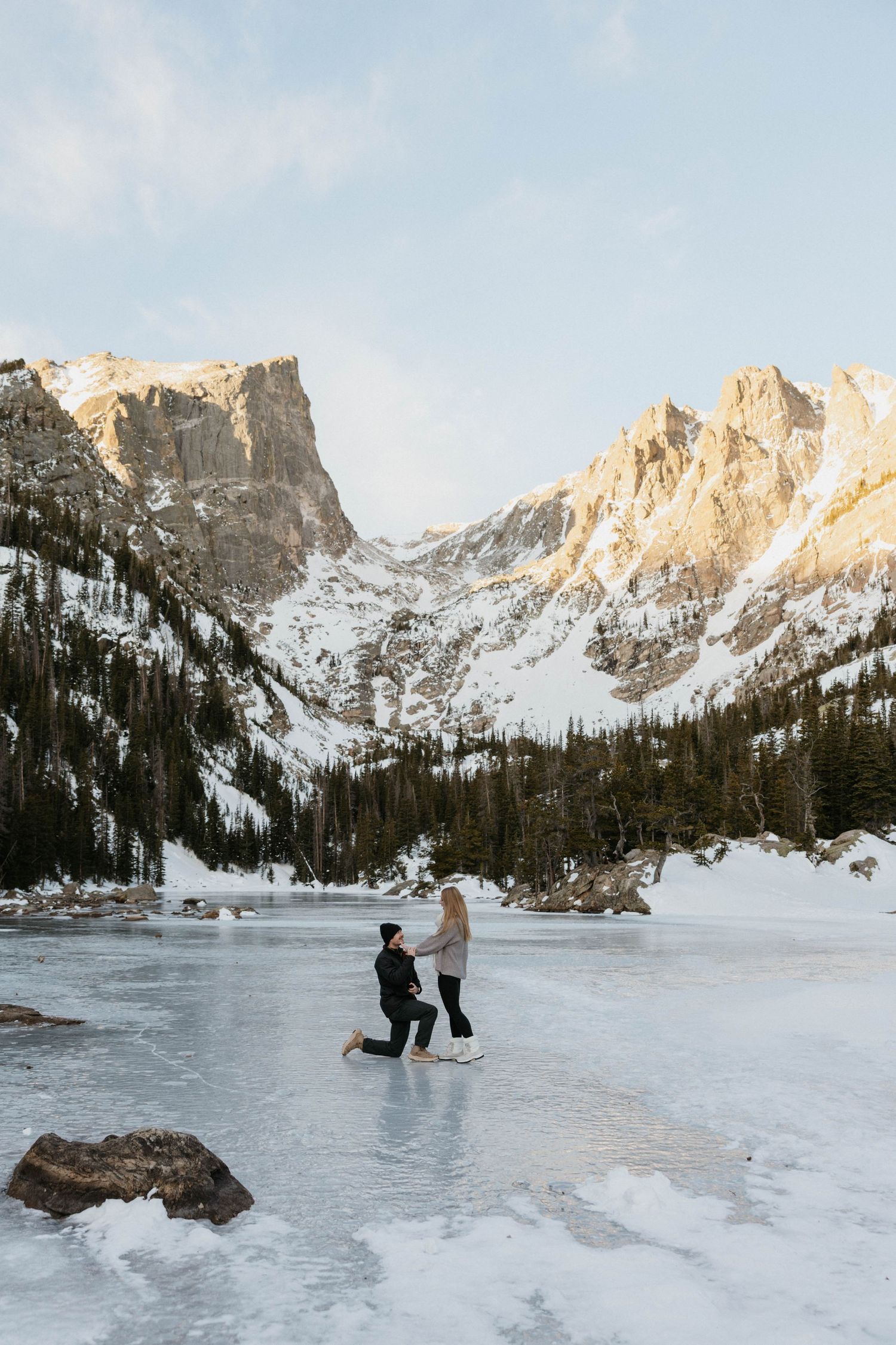 Man proposes on one knee on a frozen mountain lake surrounded by snow-capped peaks and pine trees in winter.