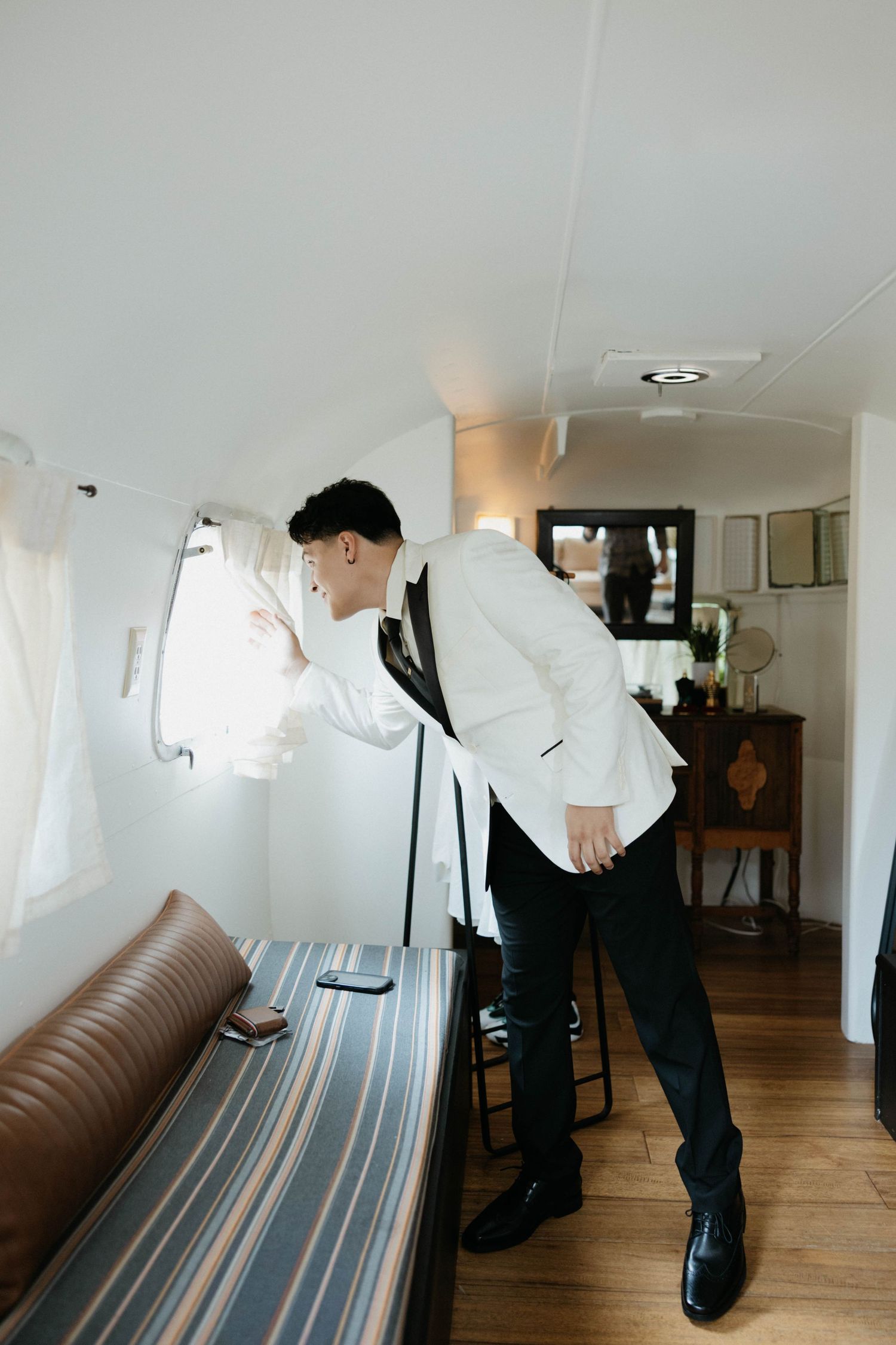 A groom in a white tuxedo jacket and black pants looks out a window inside a vintage Airstream trailer.
